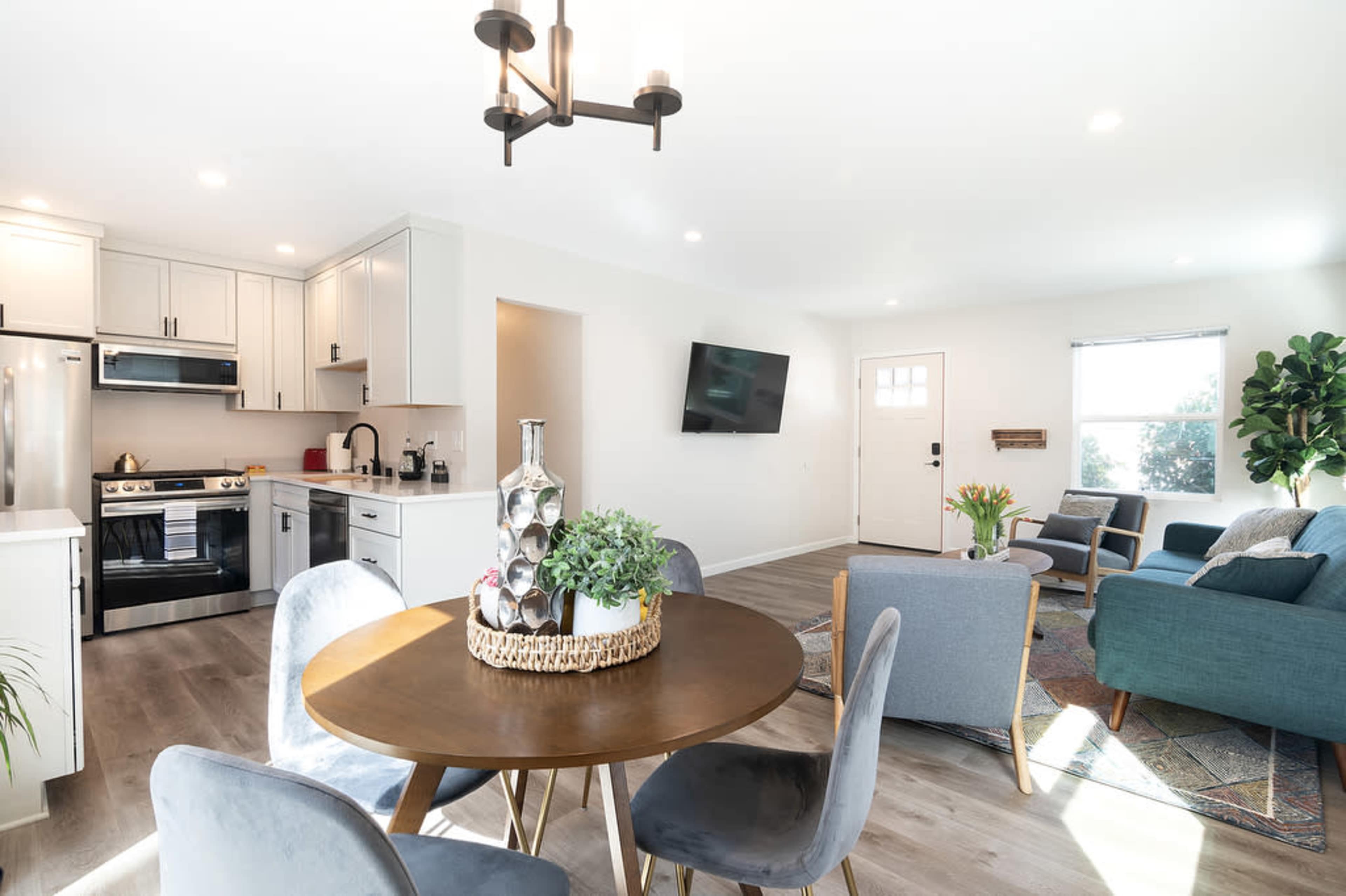 A modern living space features a dining area with a round table, four chairs, a kitchen with stainless steel appliances, and a cozy seating area with a television and indoor plants.