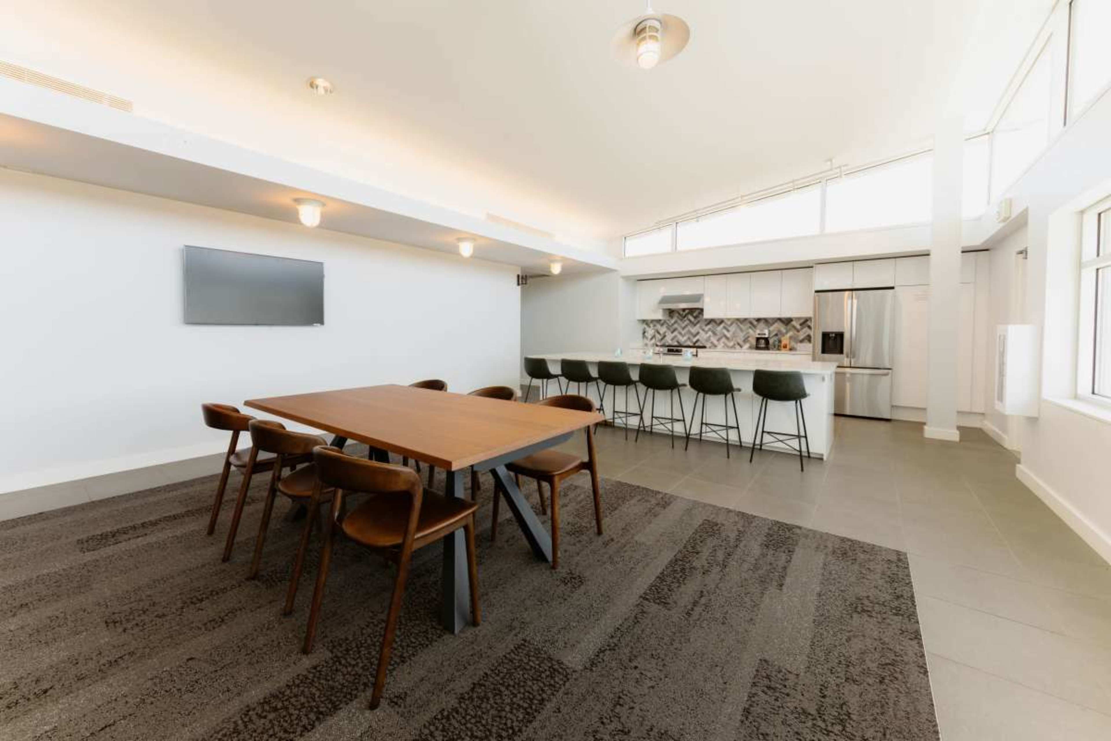 The image features a modern dining area with a large wooden table surrounded by chairs, adjacent to a kitchen with white cabinetry and bar seating.