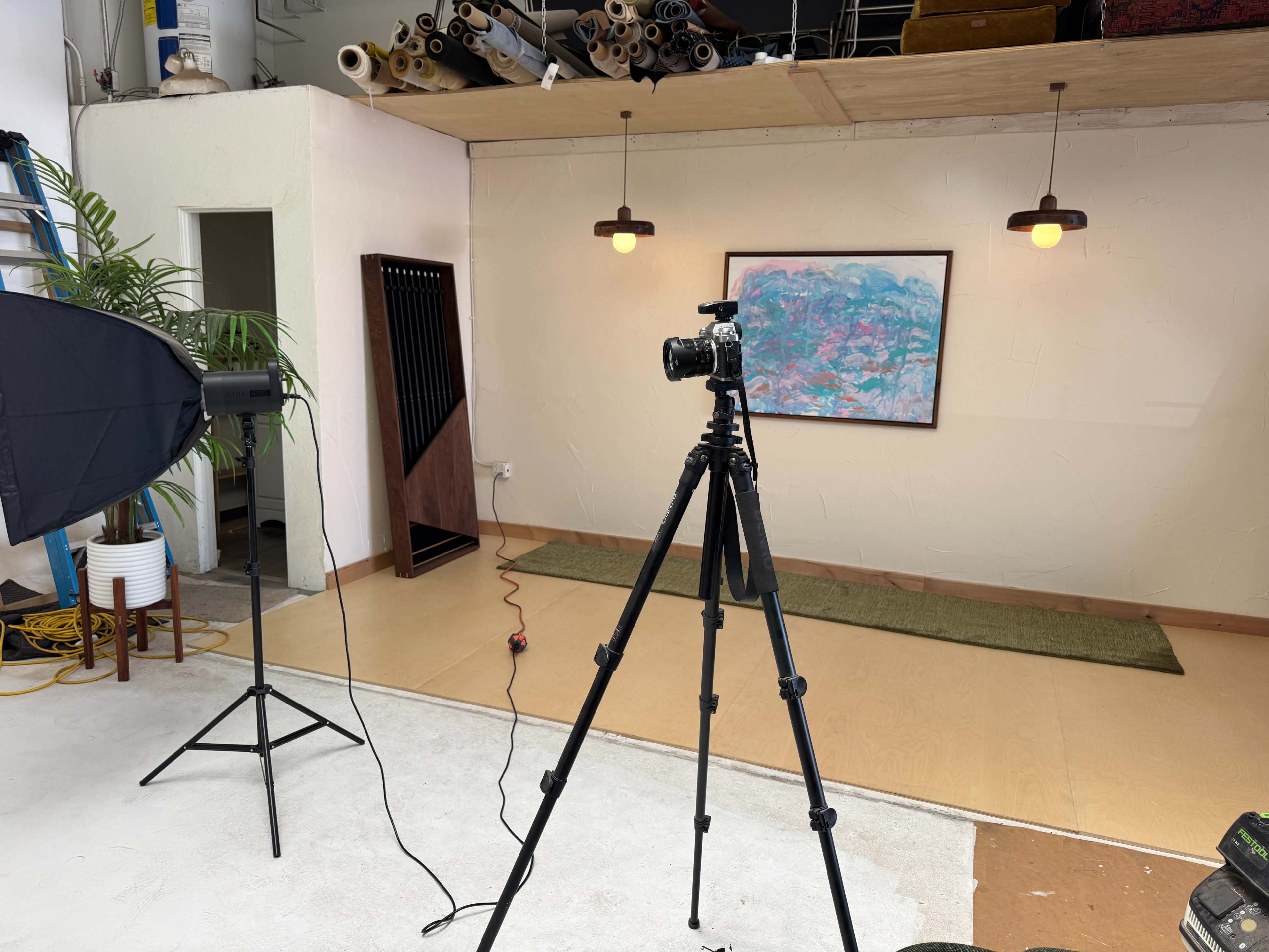 A camera on a tripod is set up in a brightly lit studio space featuring an abstract painting and pendant lights.