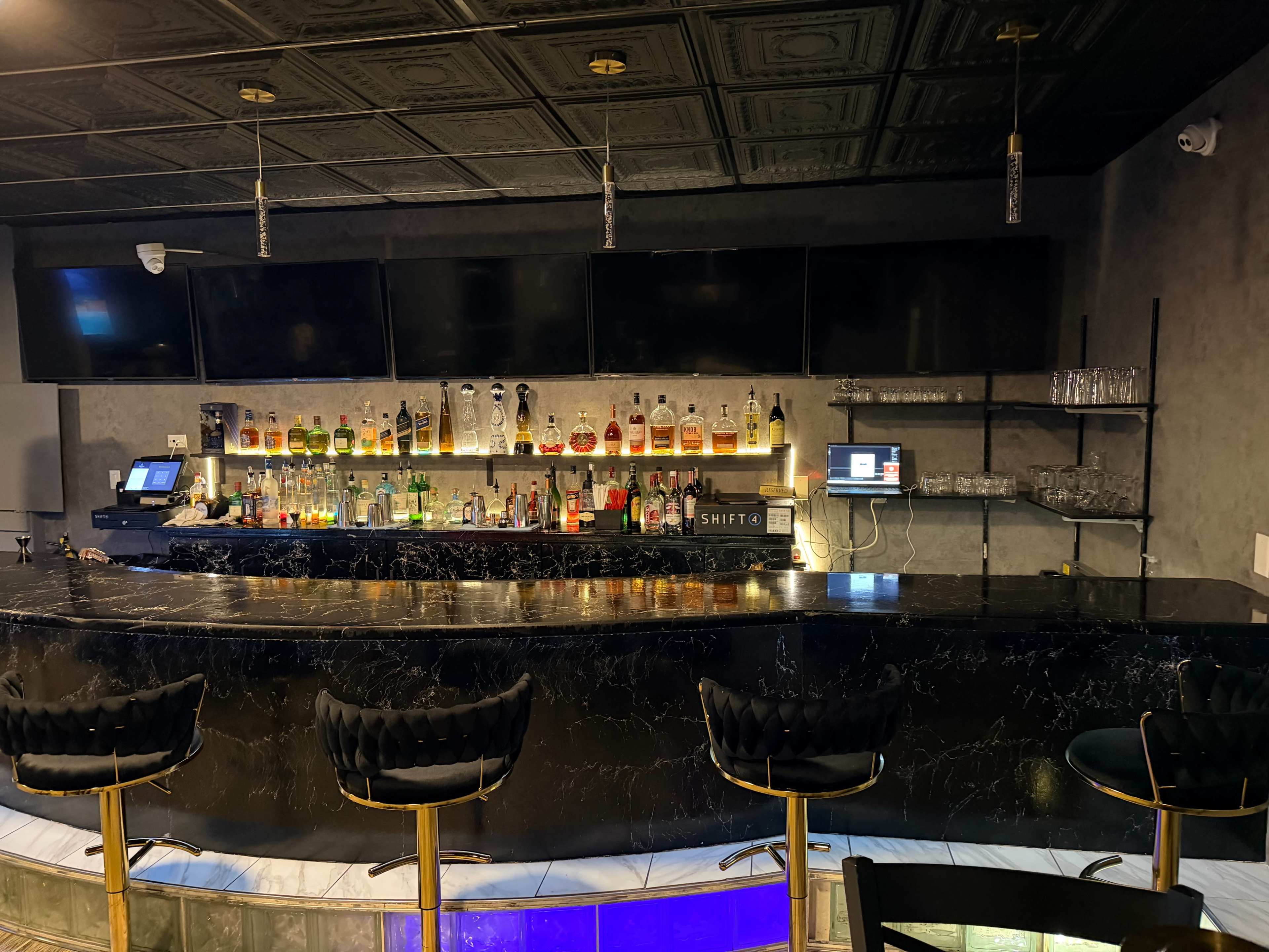 The image shows a modern bar with a marble countertop, multiple shelves displaying various bottles of alcohol, and several empty, high-backed stools.