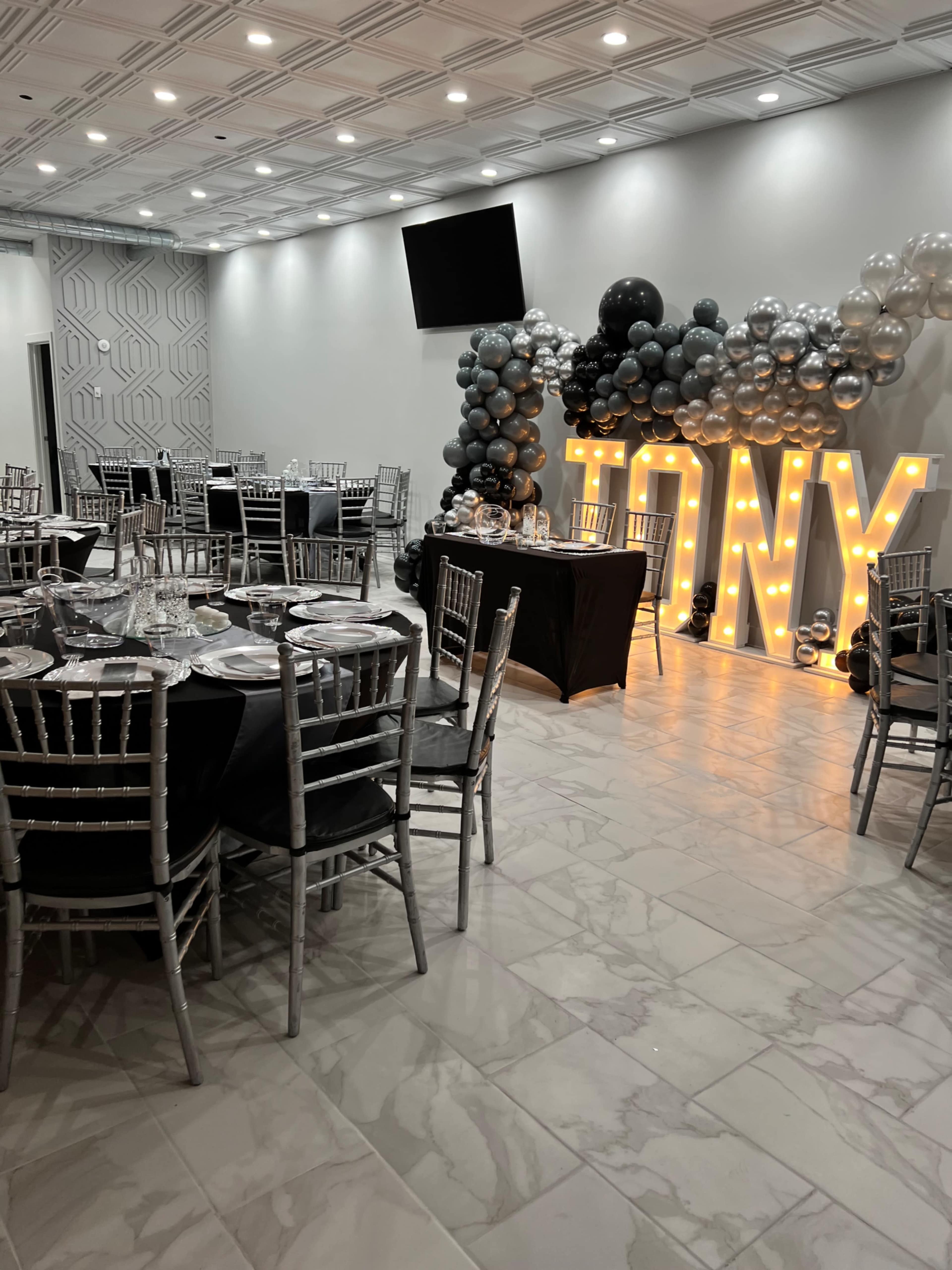 A decorated event space with tables set for a celebration, featuring silver and black balloons and illuminated letters spelling "TONY."