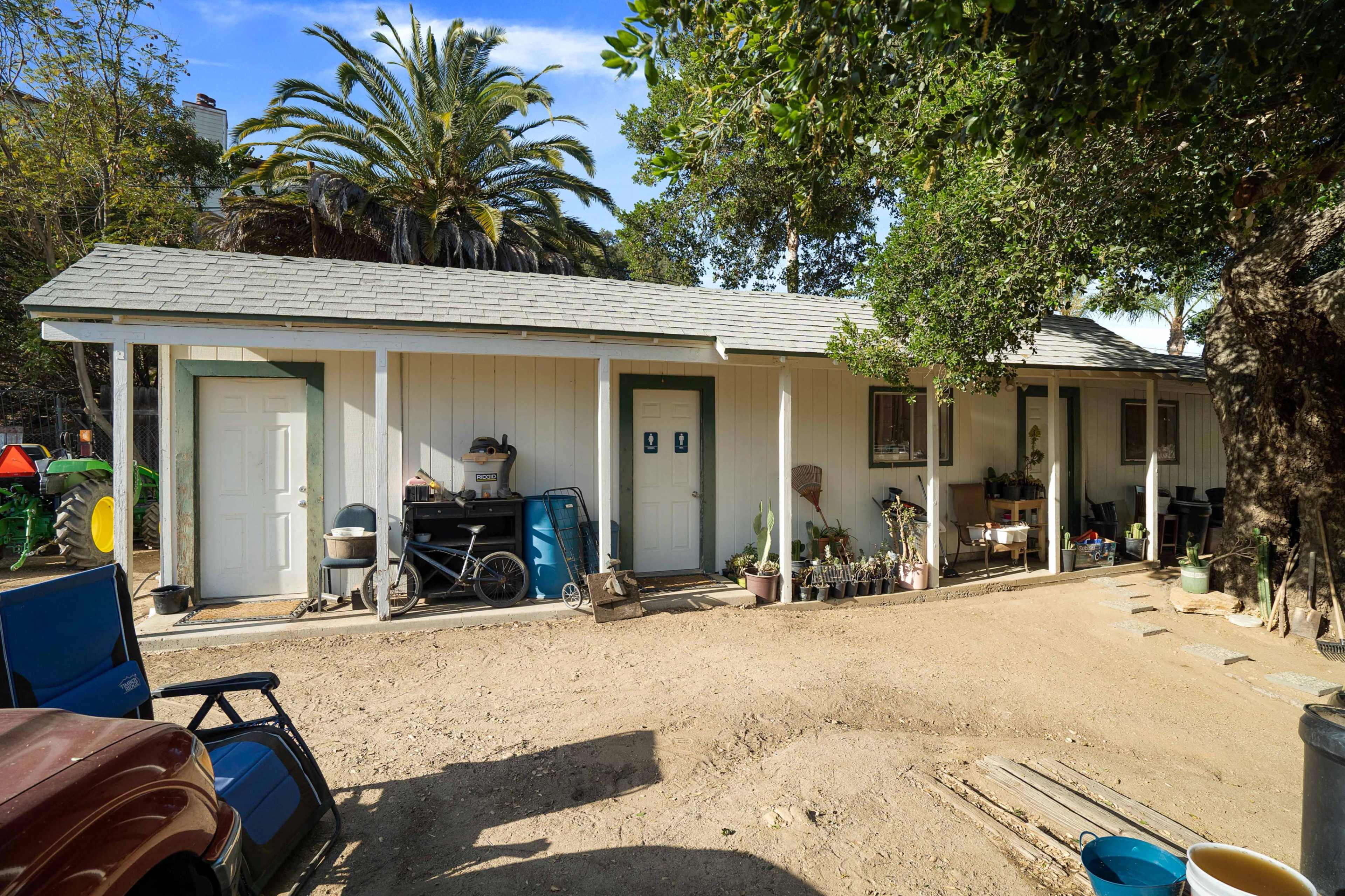 The image shows a small, single-story white building with a grey roof, surrounded by dirt and greenery, including a large palm tree.