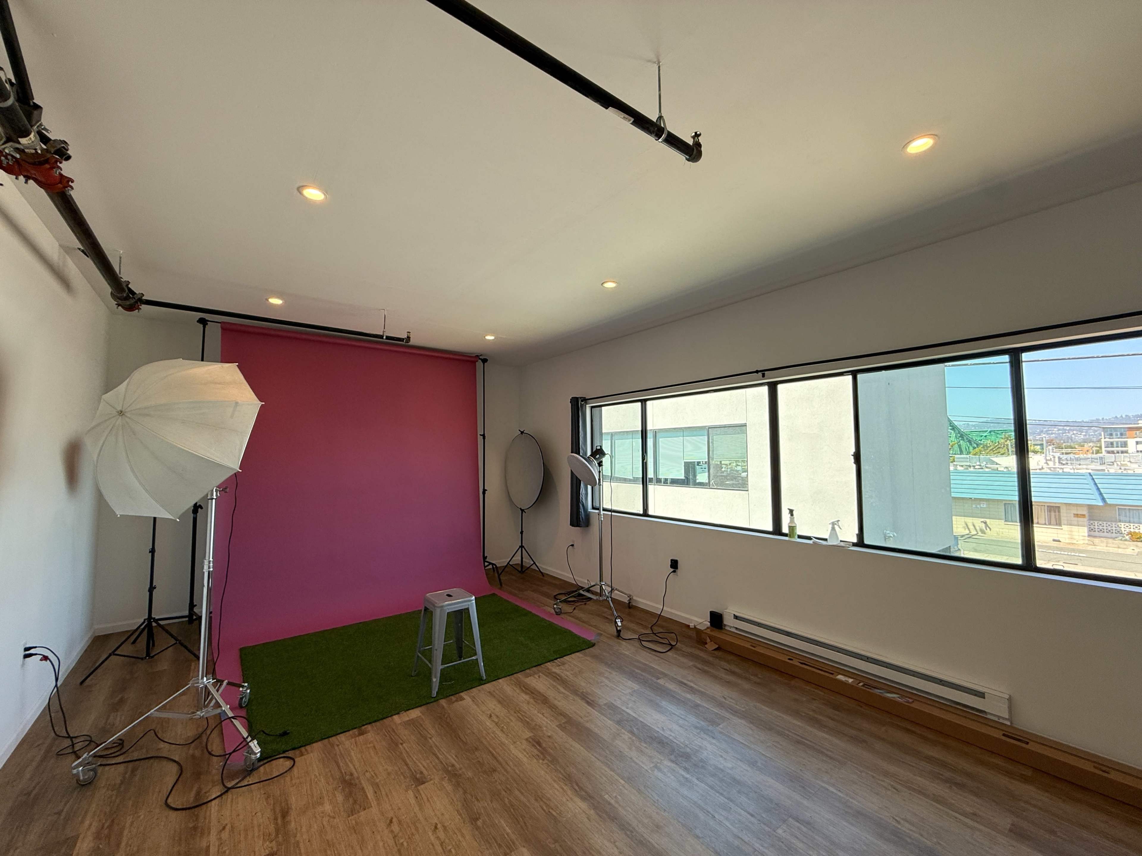 The image shows a photography studio with a pink backdrop, a stool, and lighting equipment, all set on wooden flooring.