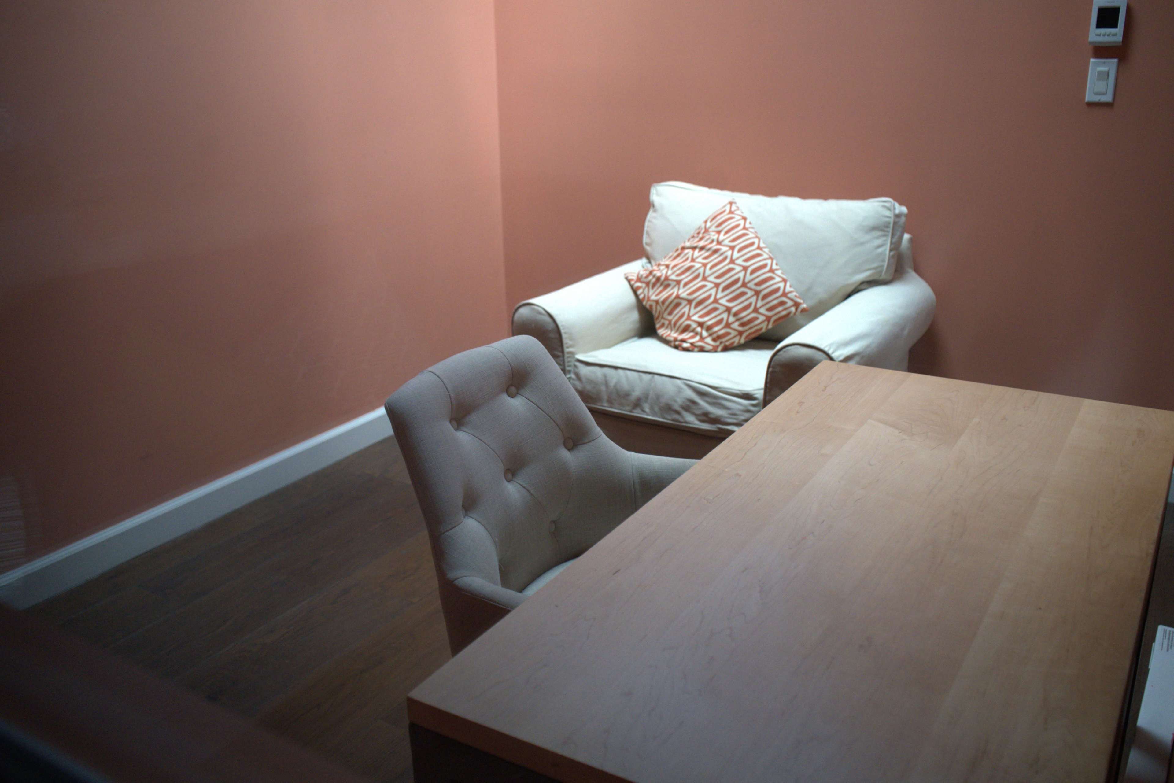 The image shows a small room with a wooden table, a chair, and a white armchair with a patterned cushion against a peach-colored wall.