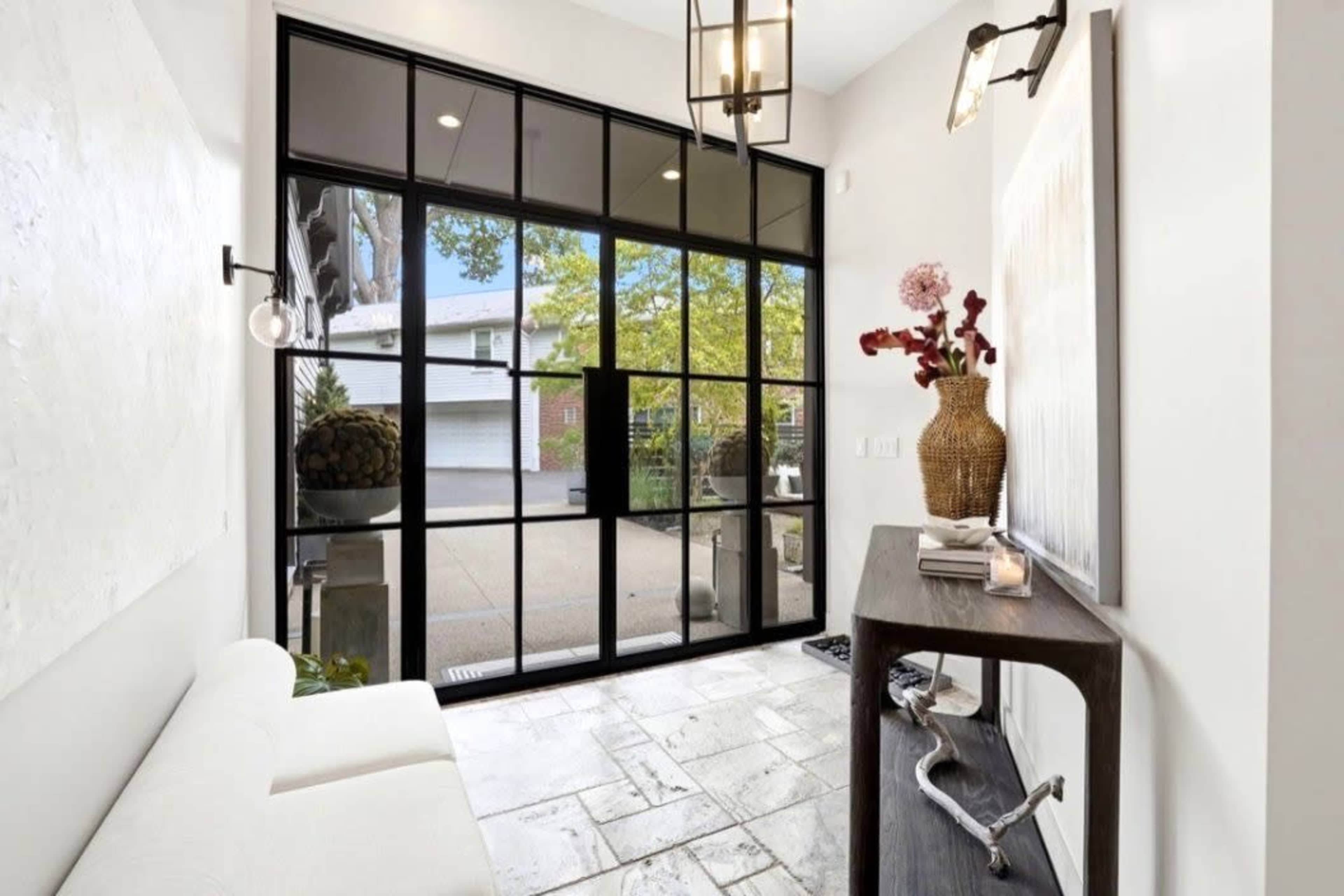 The image shows a modern entryway featuring a large glass door with black frames, a stone tile floor, a small console table, and decorative plants.