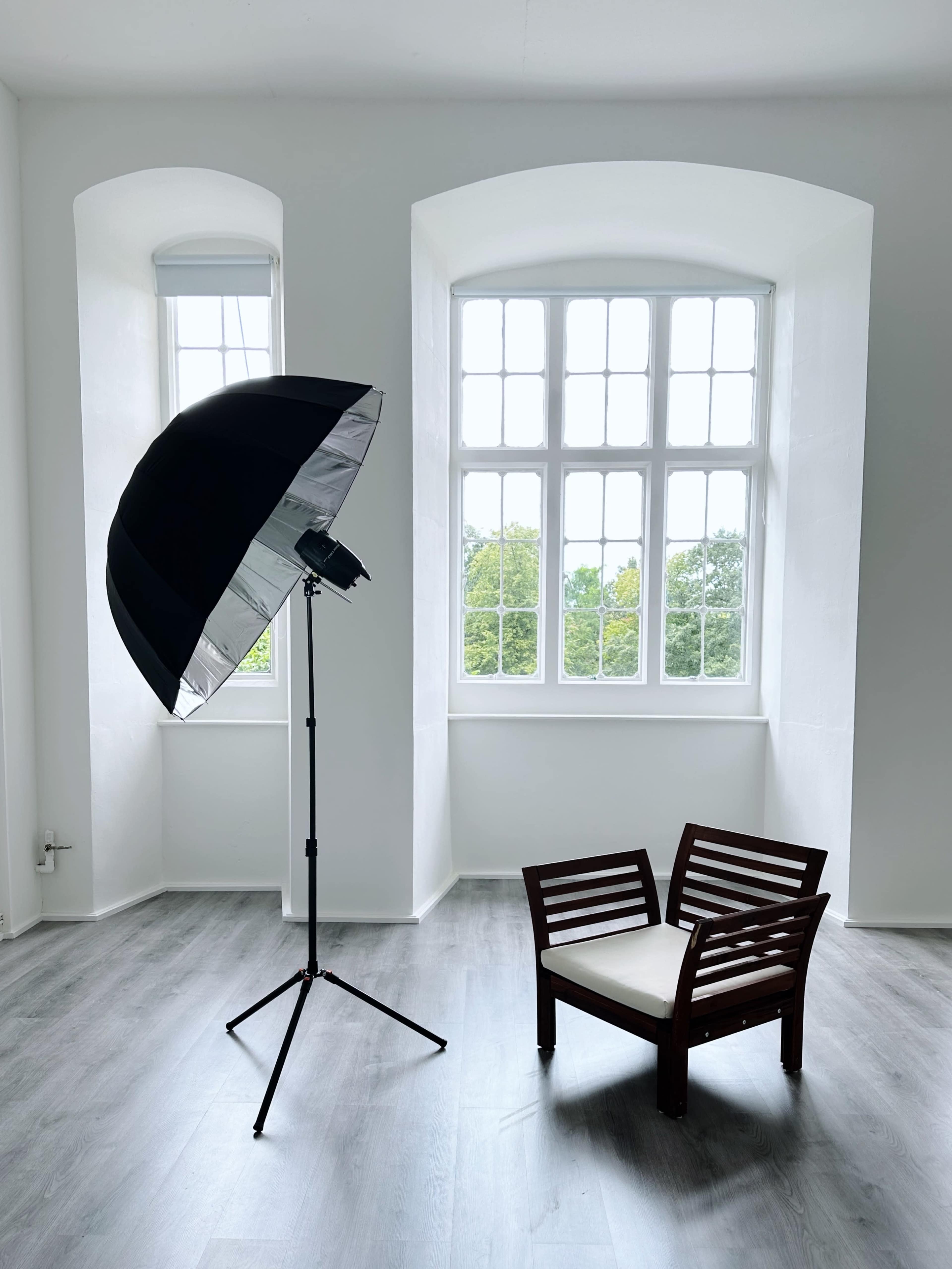 A large umbrella light stands next to a wooden chair in a bright, minimalist room with tall windows.