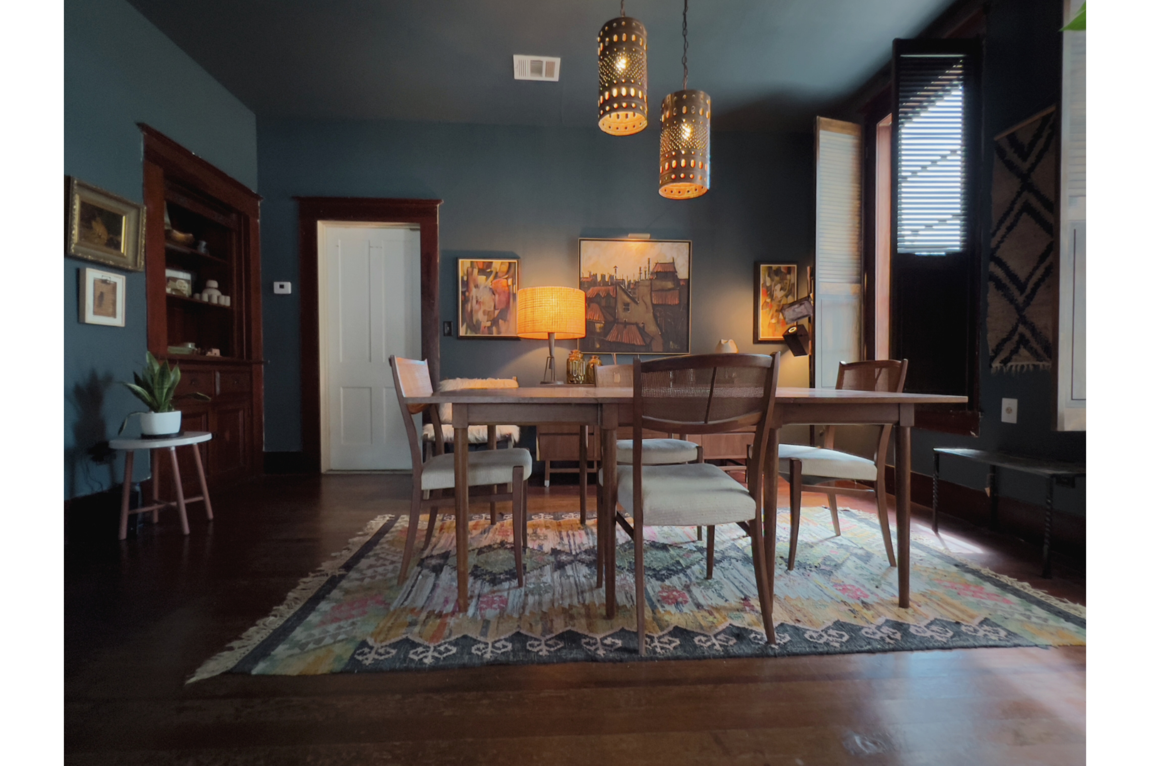 The image depicts a dining room with a wooden table surrounded by chairs, illuminated by pendant lights, with artwork on the walls and a rug on the floor.