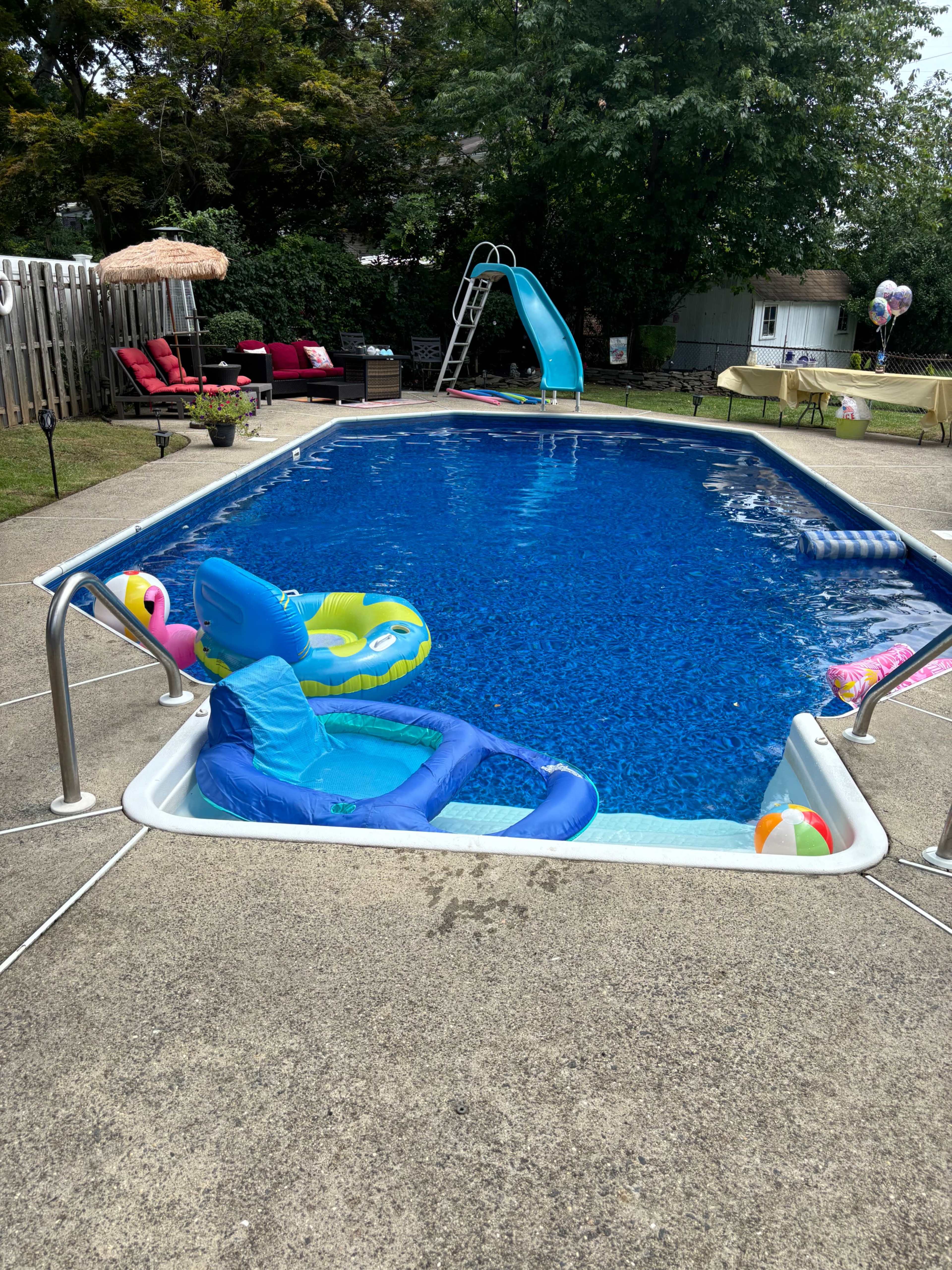A swimming pool with a blue slide, inflatable pool floats, and lounge chairs is surrounded by a concrete deck and greenery.