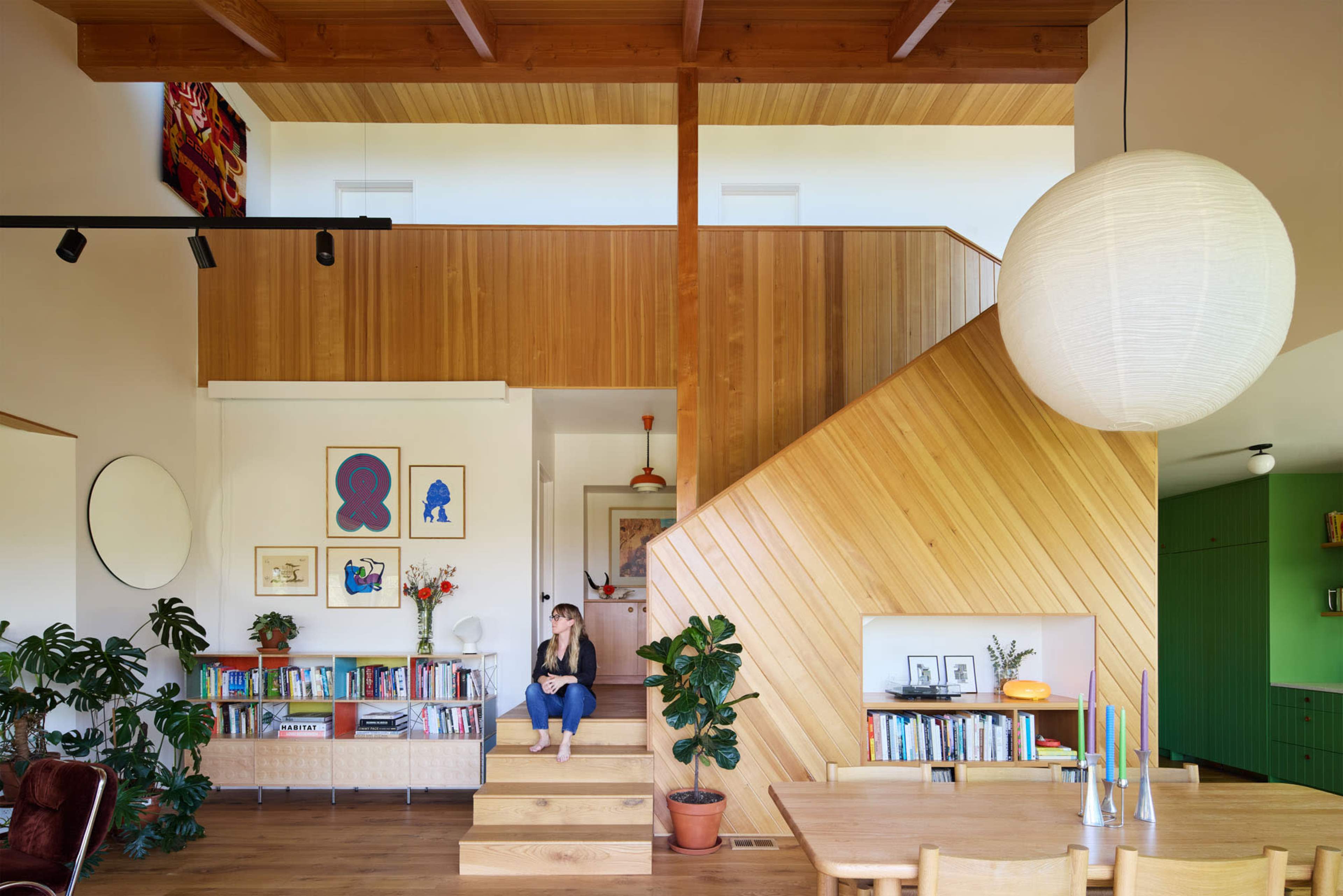 A woman sits on staircase steps in a modern, wooden interior adorned with art and plants.