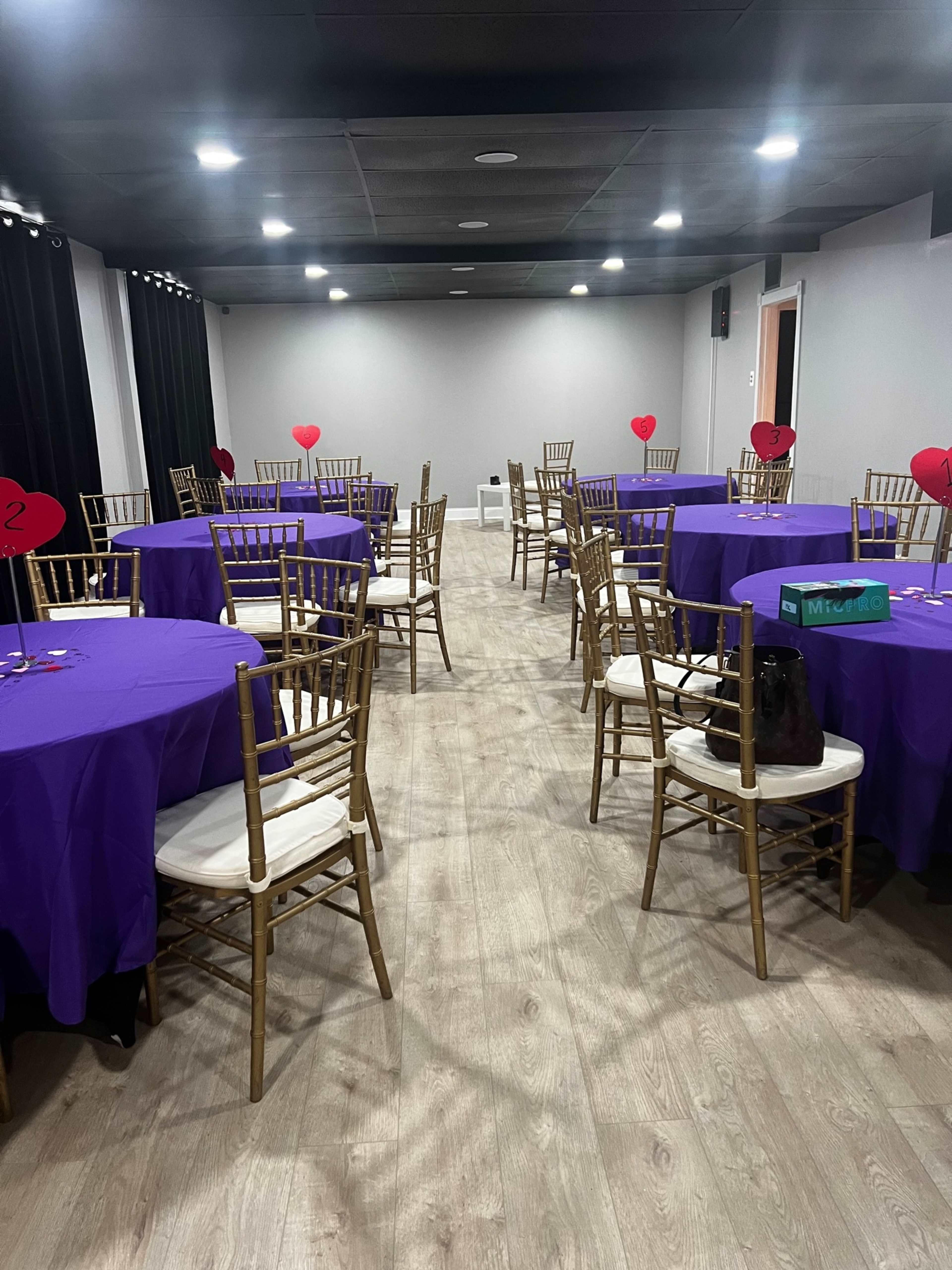 A banquet hall set up with several round tables covered in purple cloths, each surrounded by gold chairs and decorated with heart-shaped centerpieces.