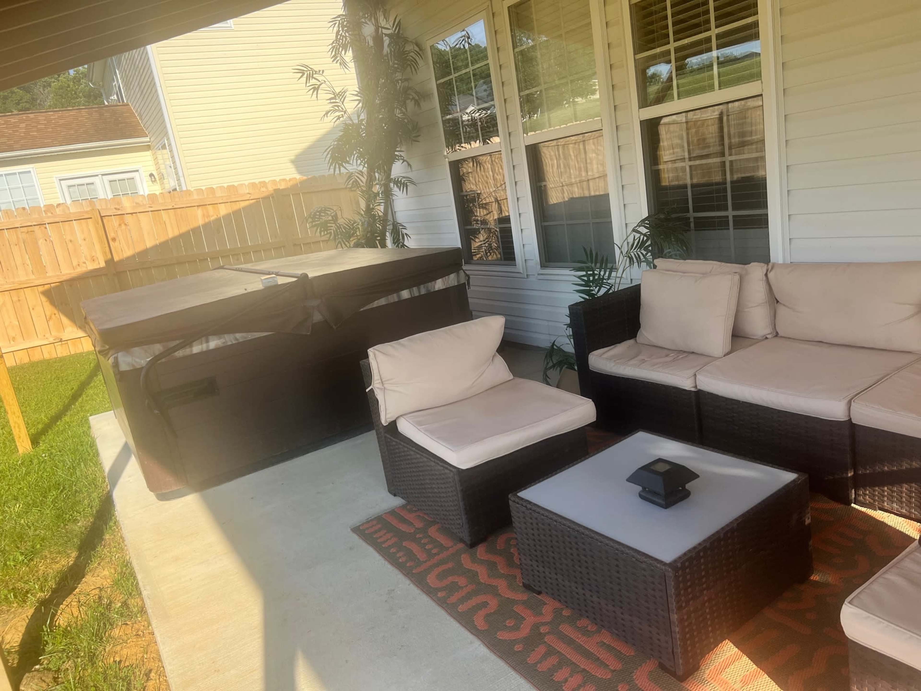 A patio with a beige sectional sofa, a small table, and a hot tub covered with a tarp, surrounded by a wooden fence and grass.