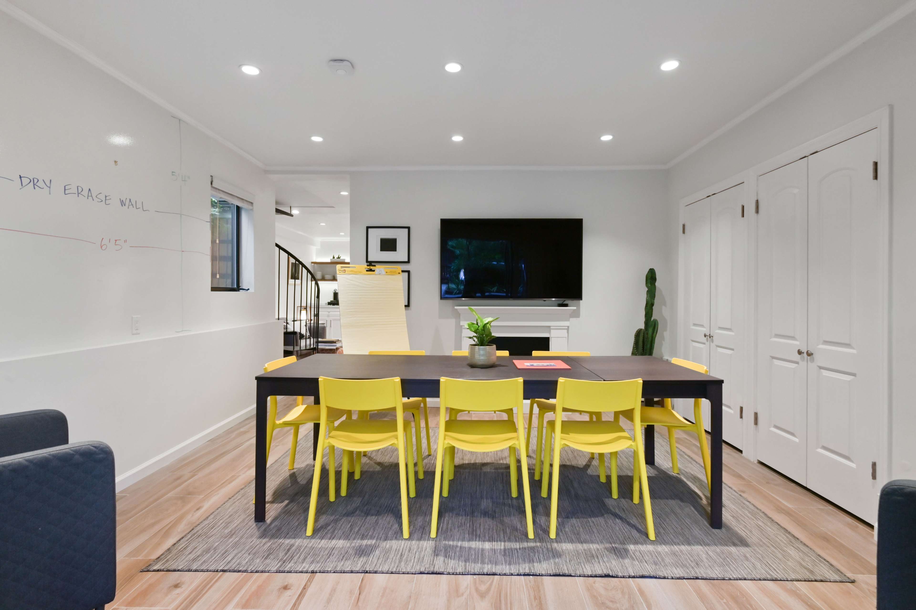 A modern meeting room features a large black table surrounded by bright yellow chairs, with a dry erase wall on one side and a television mounted above a fireplace on the opposite wall.