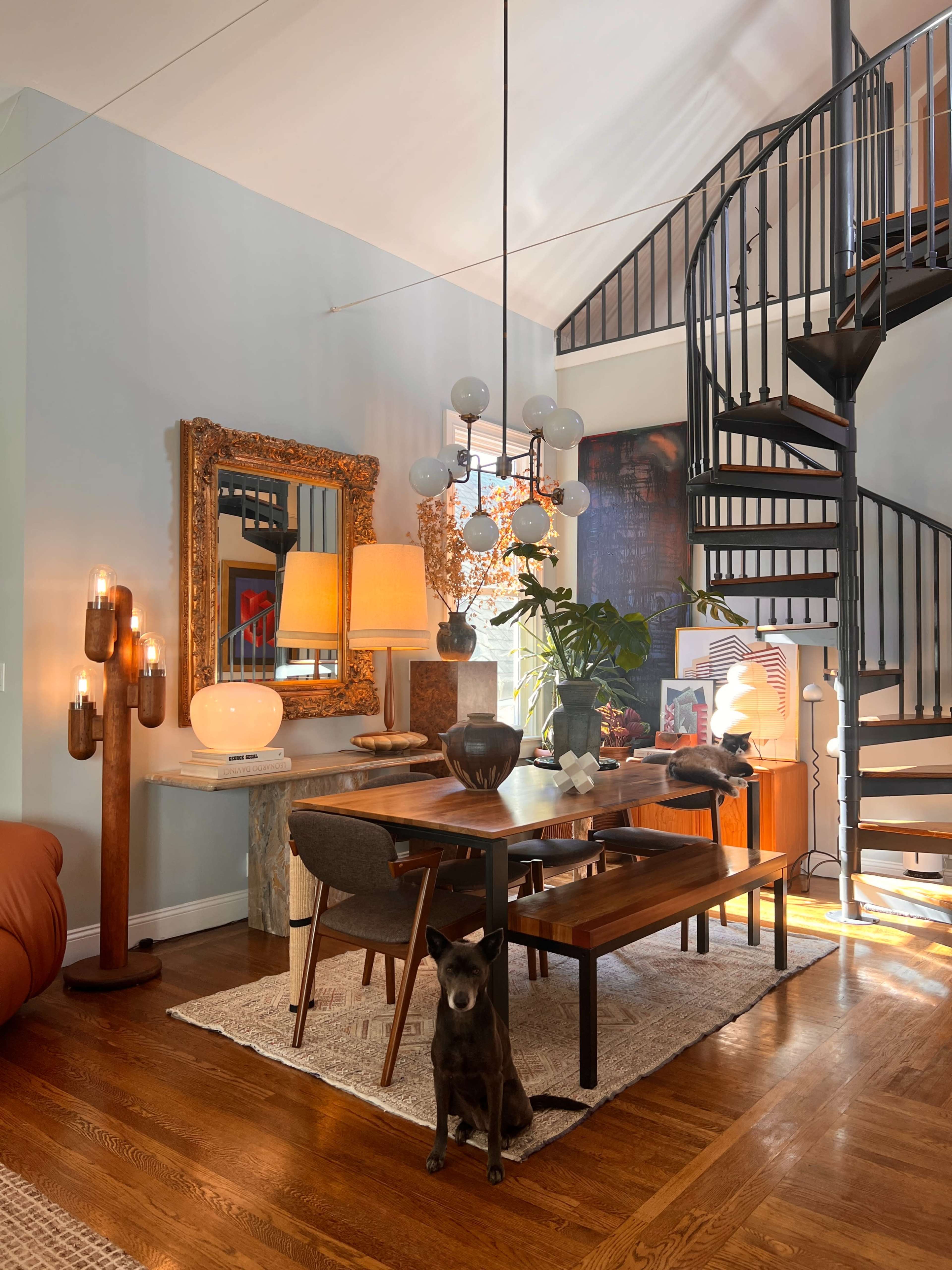 A dining area features a wooden table surrounded by chairs, with decorative lamps, a large mirror, and a spiral staircase in the background.