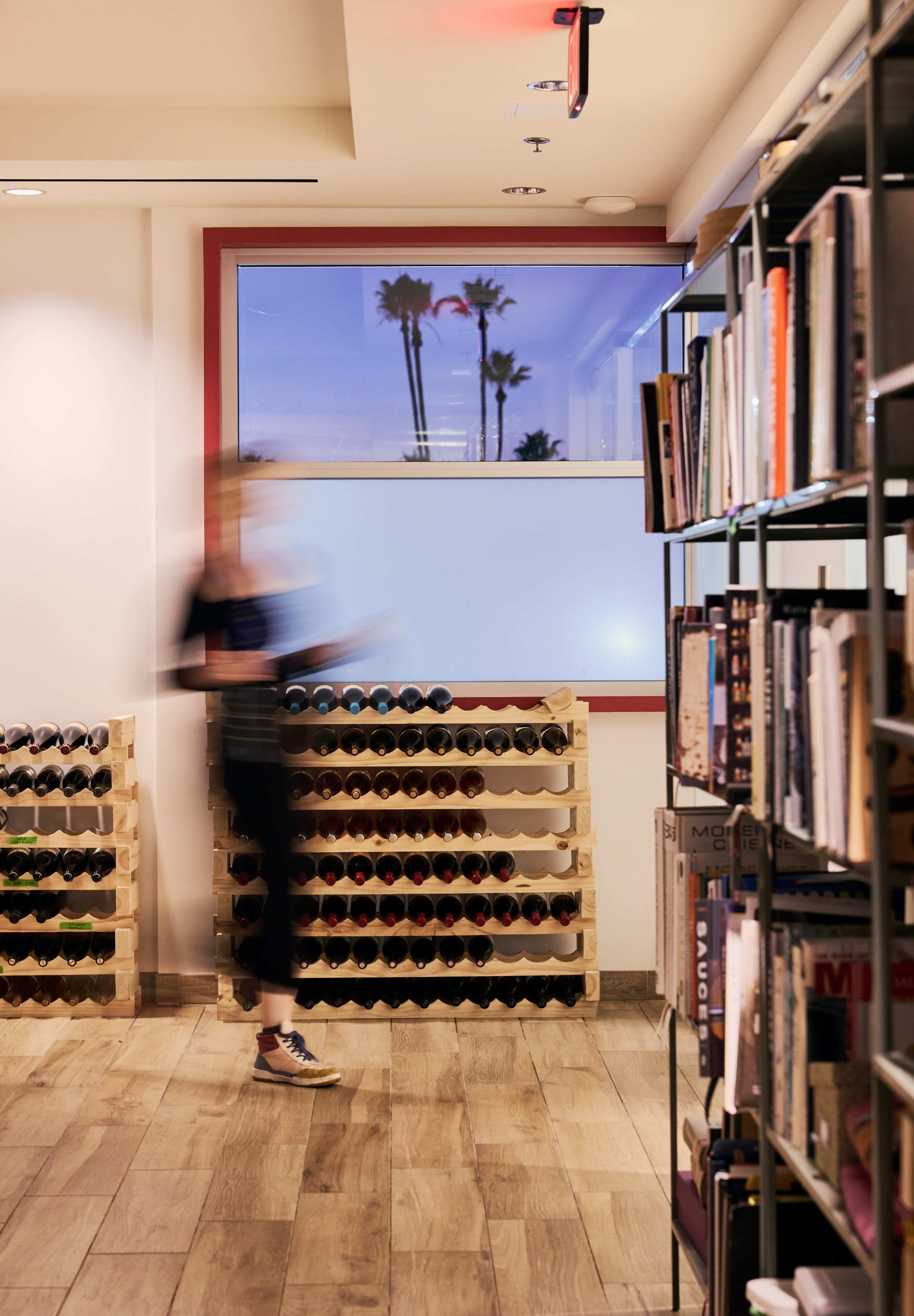 A person walks past a wine rack and bookshelves in a well-lit room with a window showing palm trees outside.