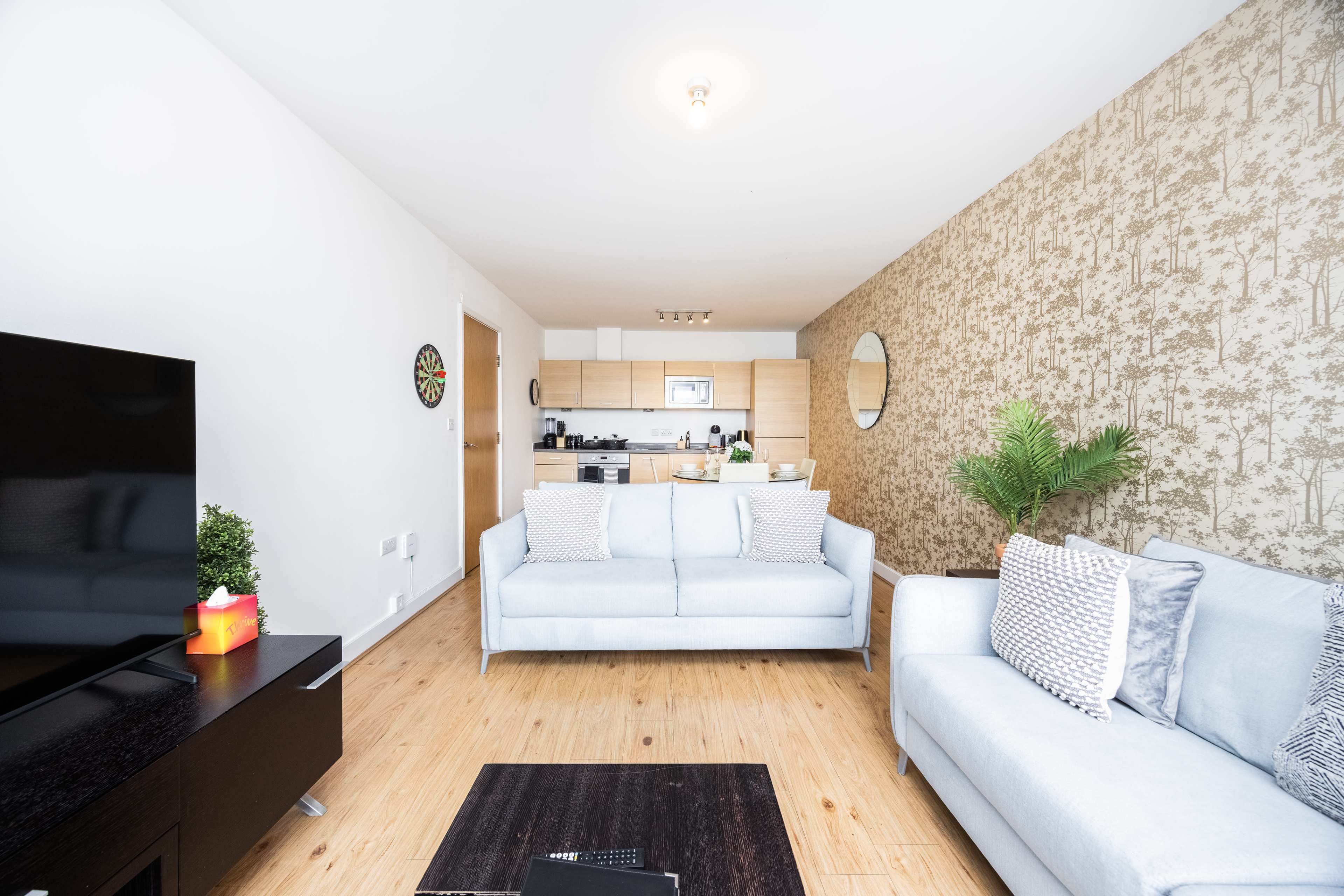 A living room features two light-colored sofas, a coffee table, and a television, with a kitchen visible in the background.