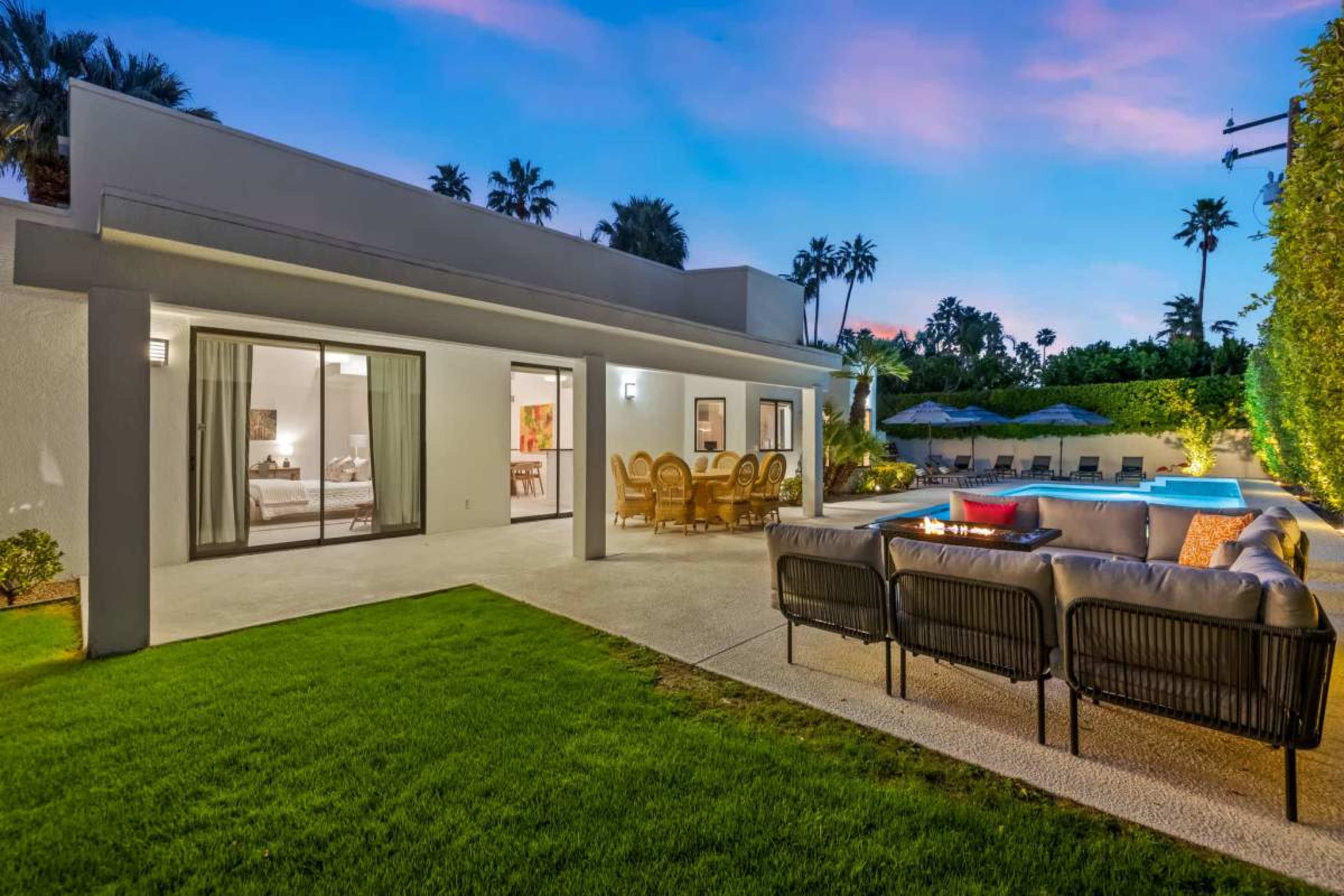 A modern house with a spacious patio and pool area features outdoor seating and dining arrangements surrounded by palm trees at dusk.