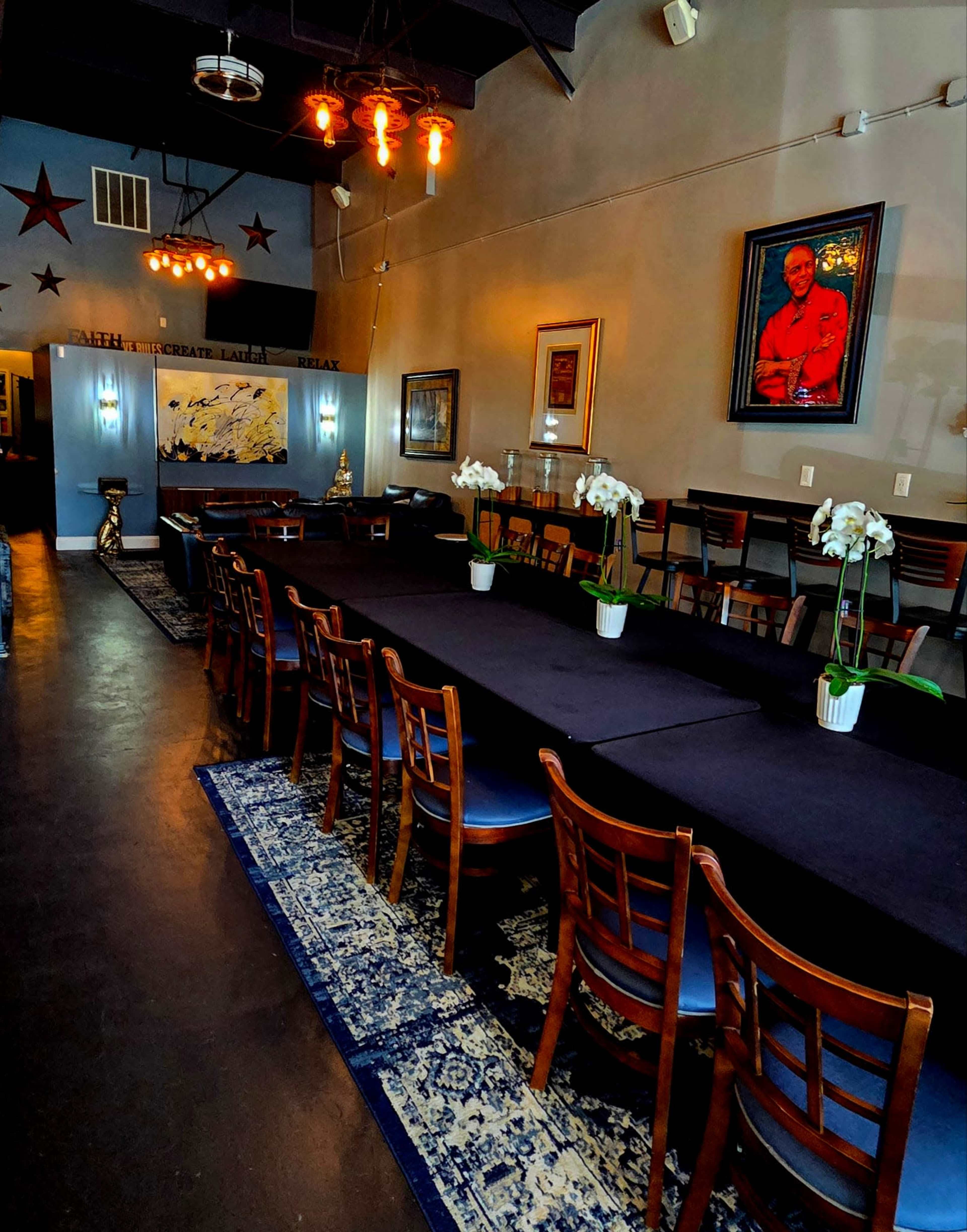 The image shows a long dining table with wooden chairs, set in a well-lit restaurant featuring artwork on the walls and a decorative rug on the floor.
