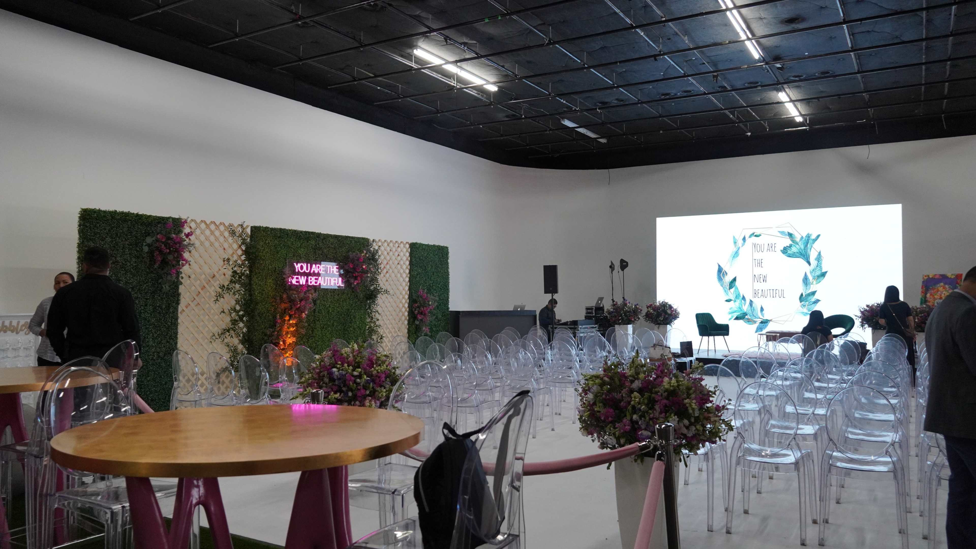 The image shows an event space with clear chairs arranged in neat rows facing a large screen and a decorated backdrop with text, alongside tables with floral centerpieces.