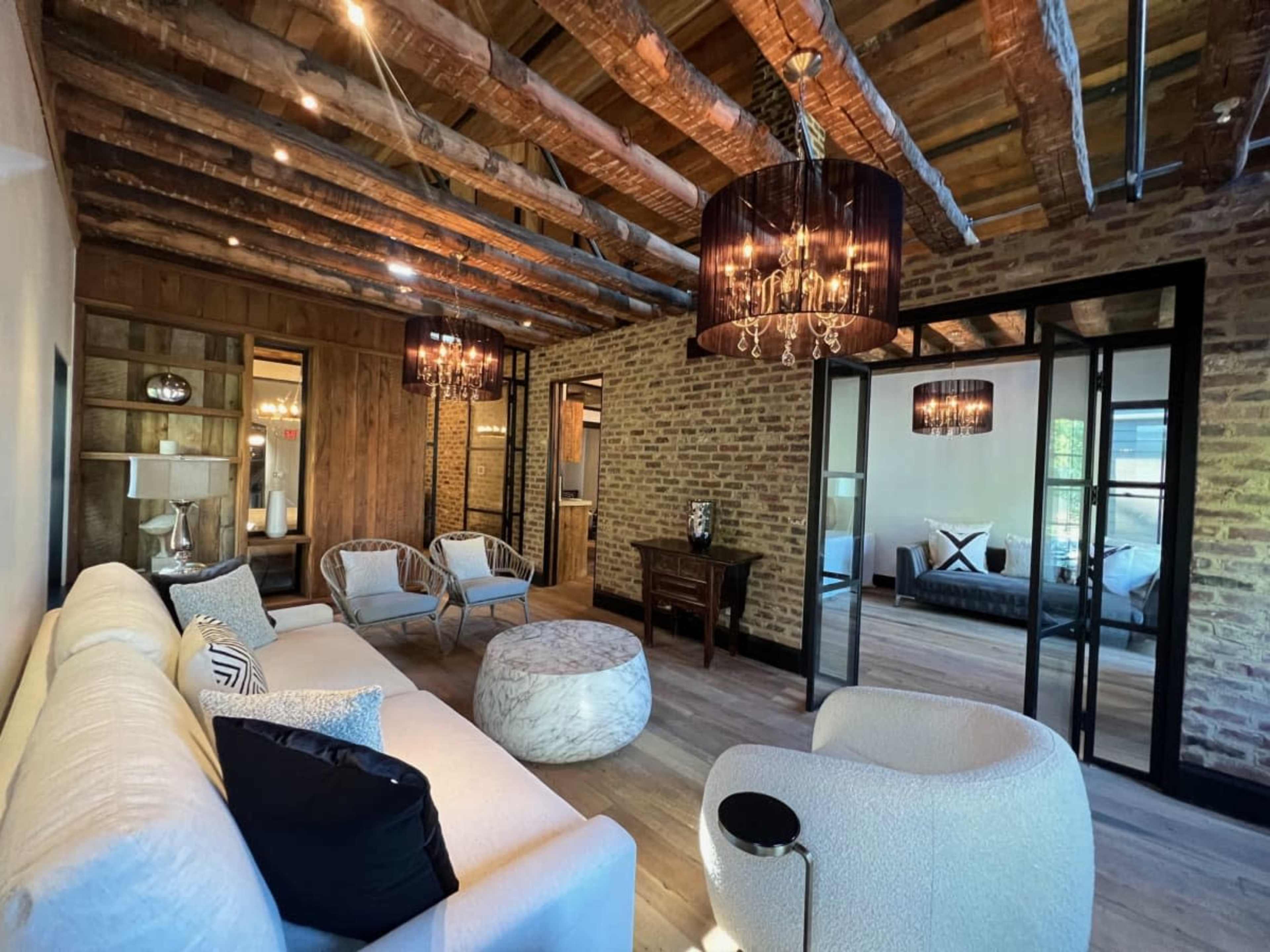 A modern living room with exposed wooden beams, brick walls, and a mix of stylish furniture, including a white sofa and light fixtures.