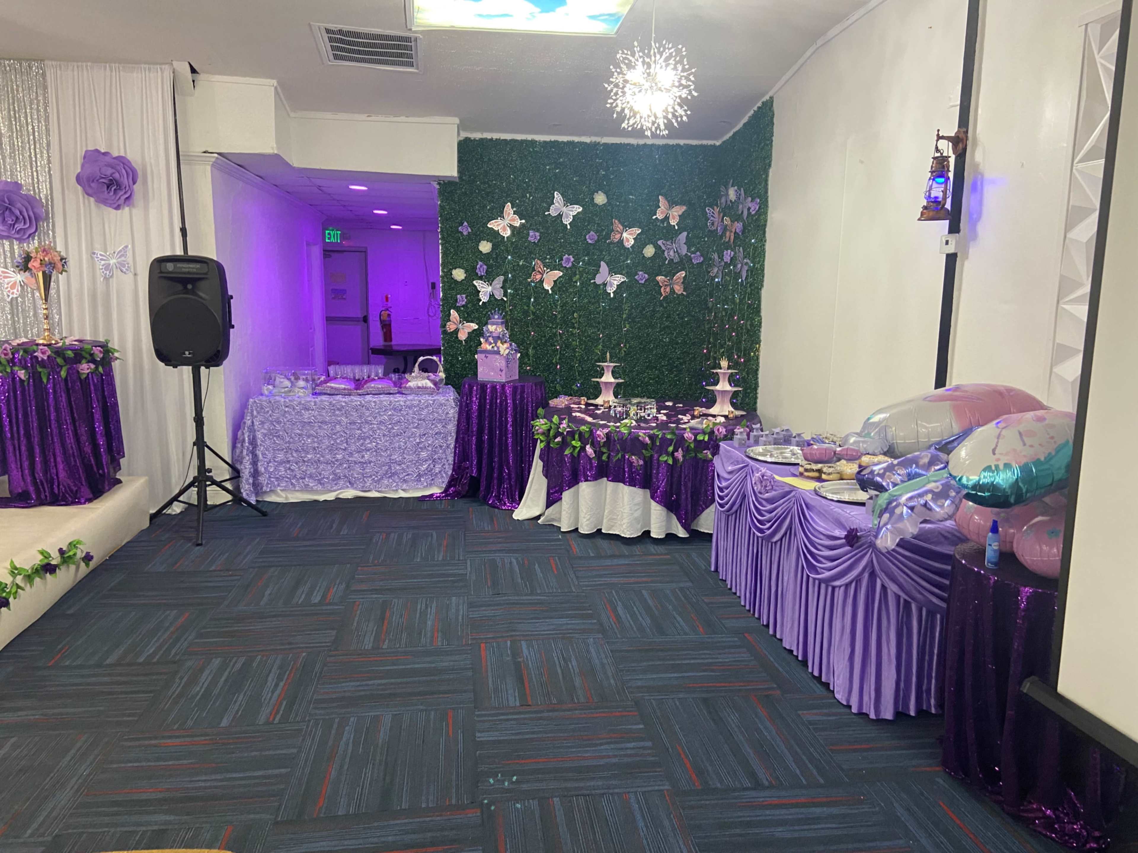 The image shows a decorated event space with purple-themed tables featuring various sweet treats and floral accents, accompanied by butterflies on the walls.
