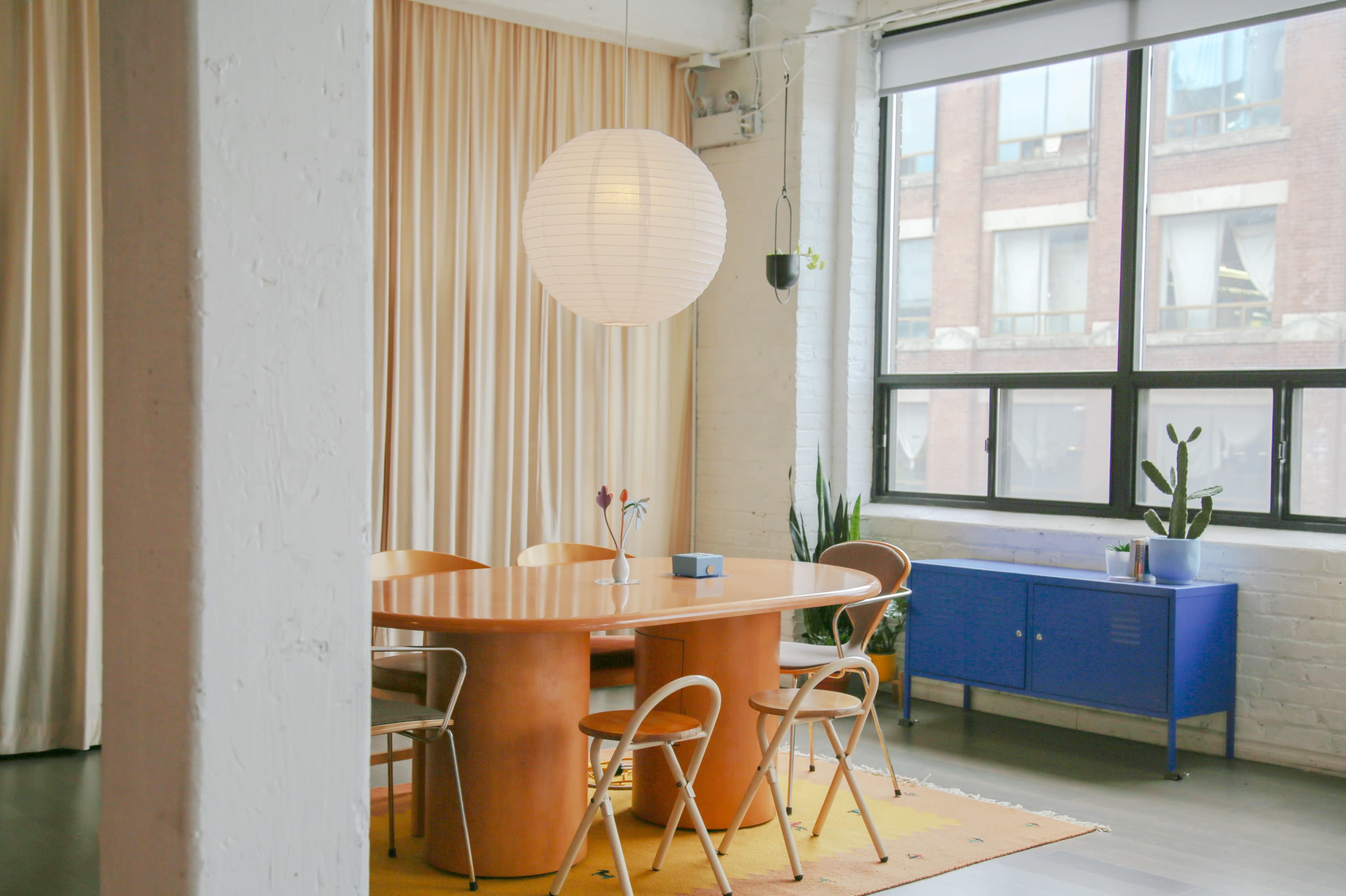 A round wooden dining table with seven chairs is positioned in a well-lit room featuring large windows and a paper lantern pendant light.