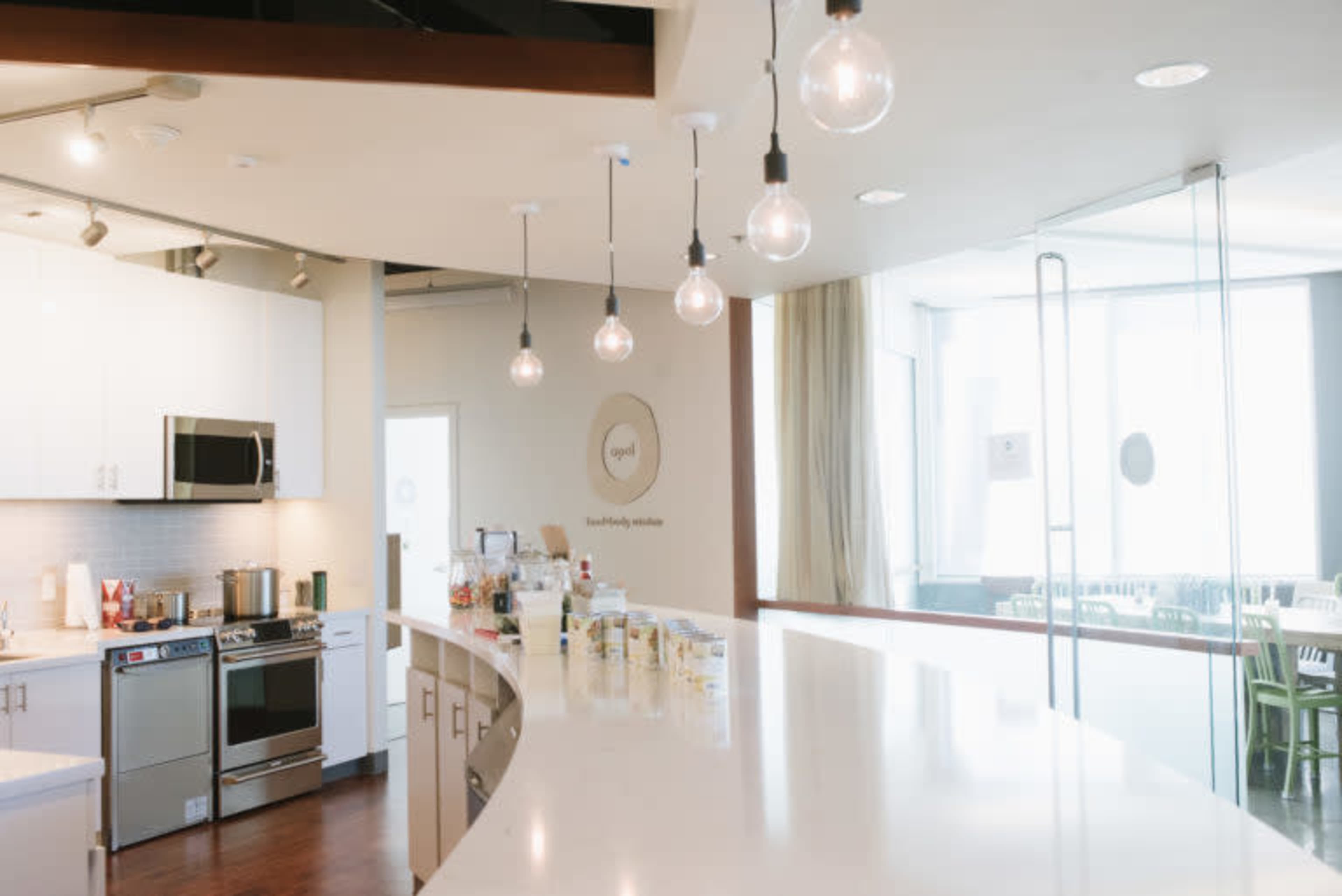 The image shows a modern kitchen with white cabinetry, stainless steel appliances, and a long counter illuminated by pendant lights.