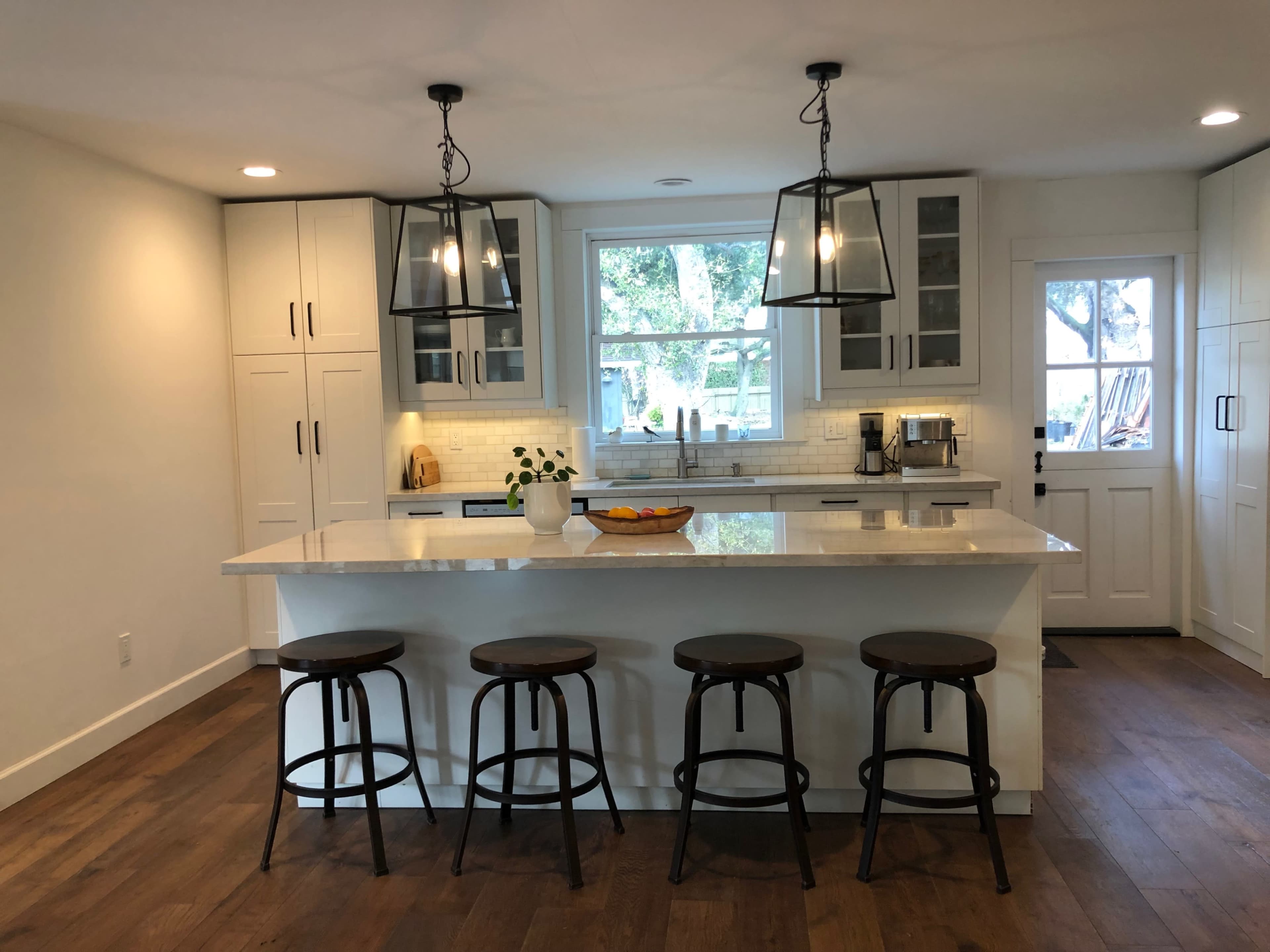 A modern kitchen features white cabinets, a large island with four wooden stools, and pendant lighting overhead.