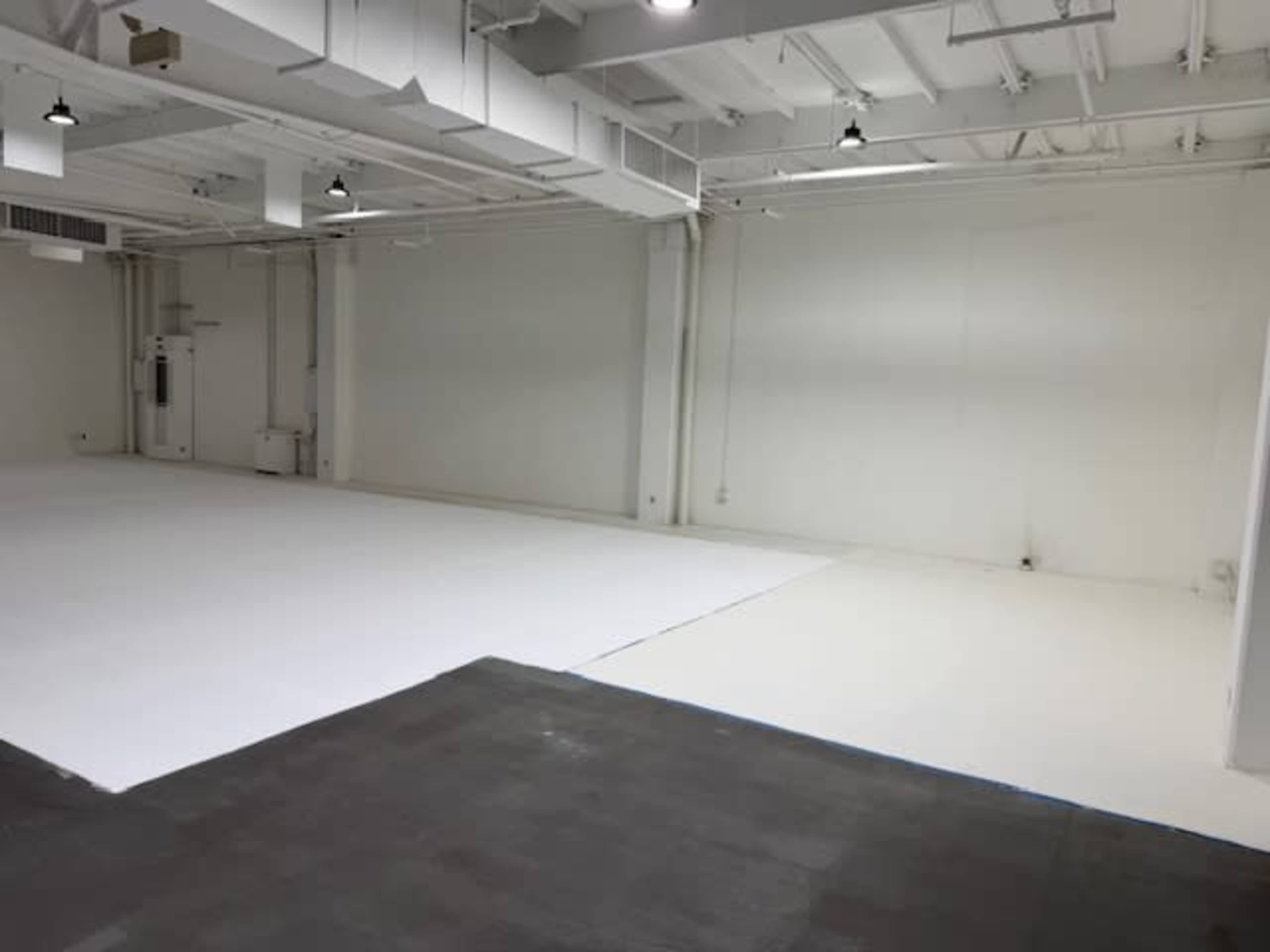The image shows a large, empty room with a white floor and plain white walls, illuminated by overhead lights.