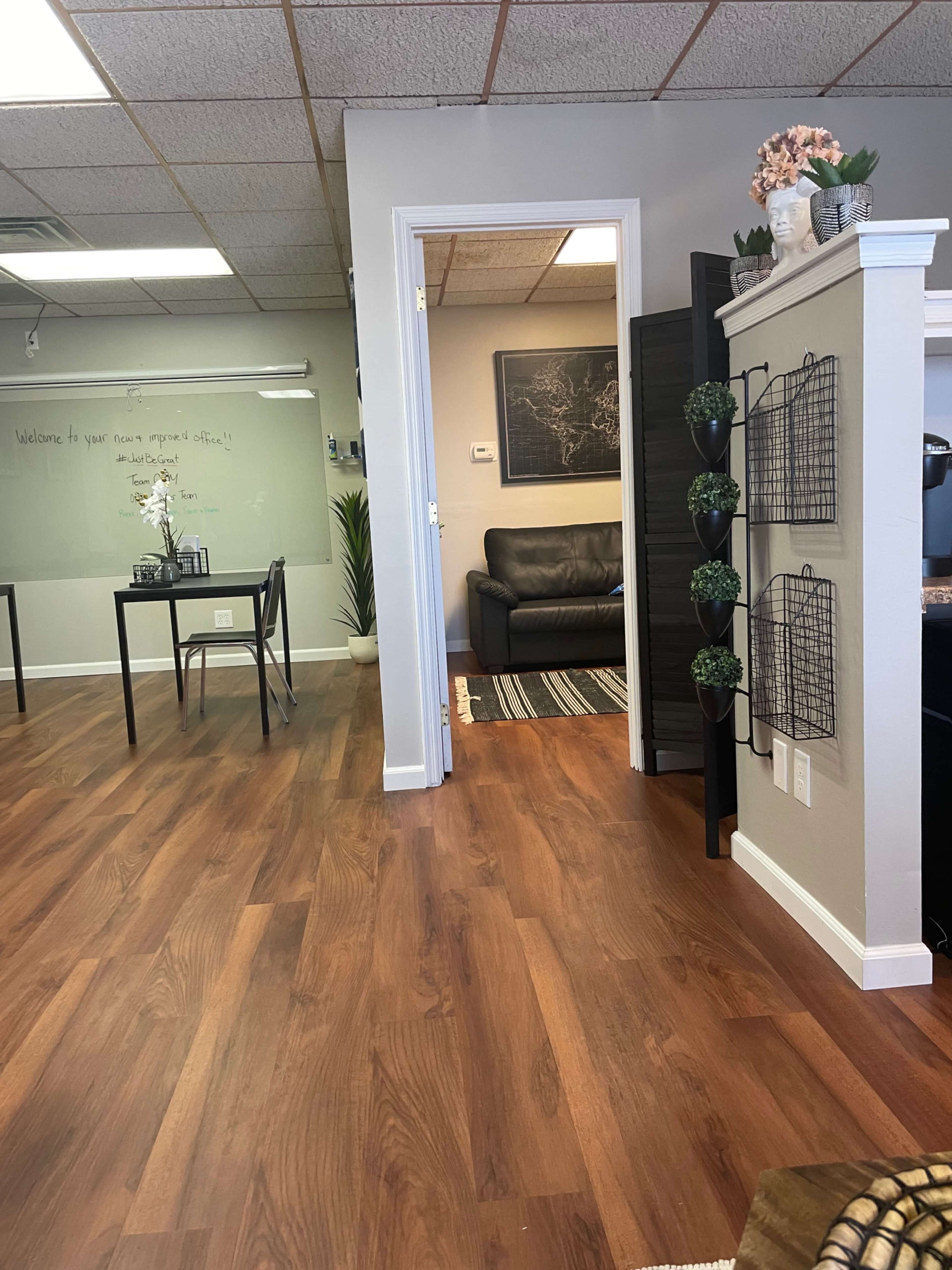 A modern office interior with wooden flooring, a reception area with a table and chairs, and a doorway leading to a lounge area with a sofa.
