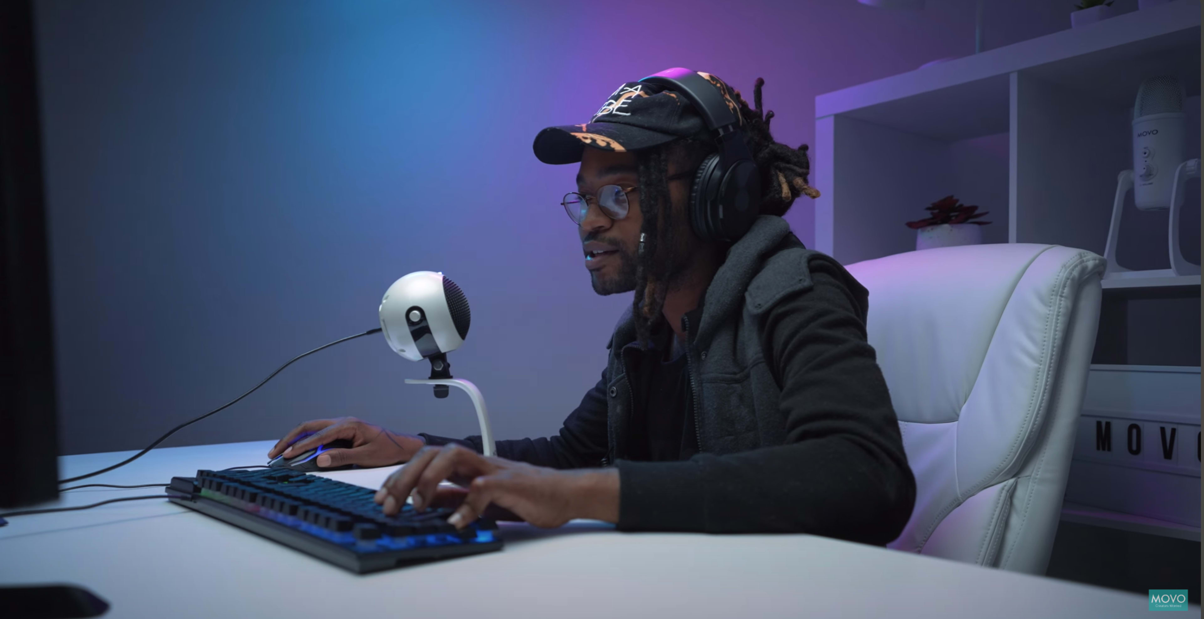 A person with headphones is gaming at a computer desk, focused on the screen with a microphone positioned nearby.