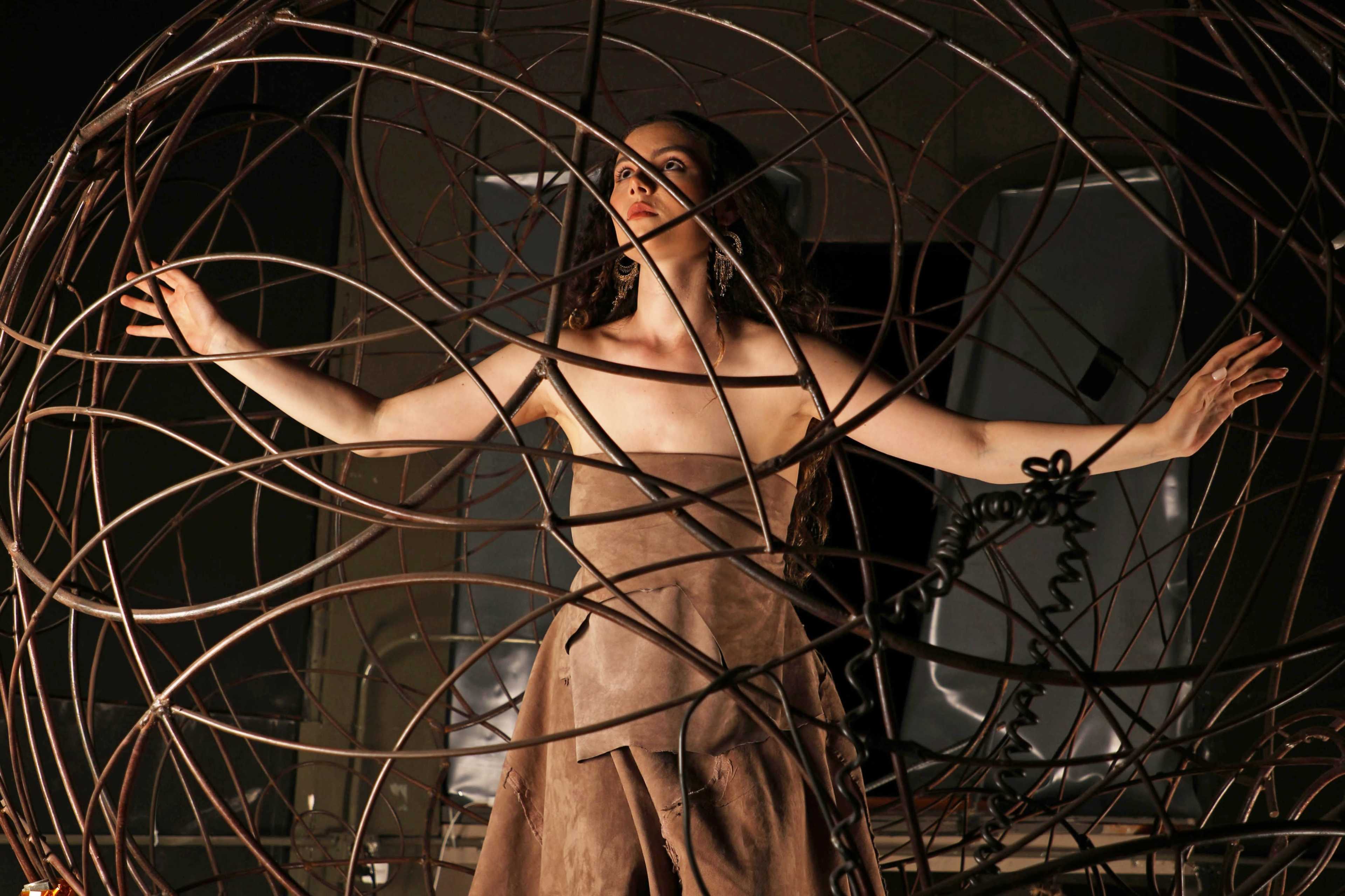 A woman in a brown dress stands inside a large, intricate wire sculpture with her arms outstretched.