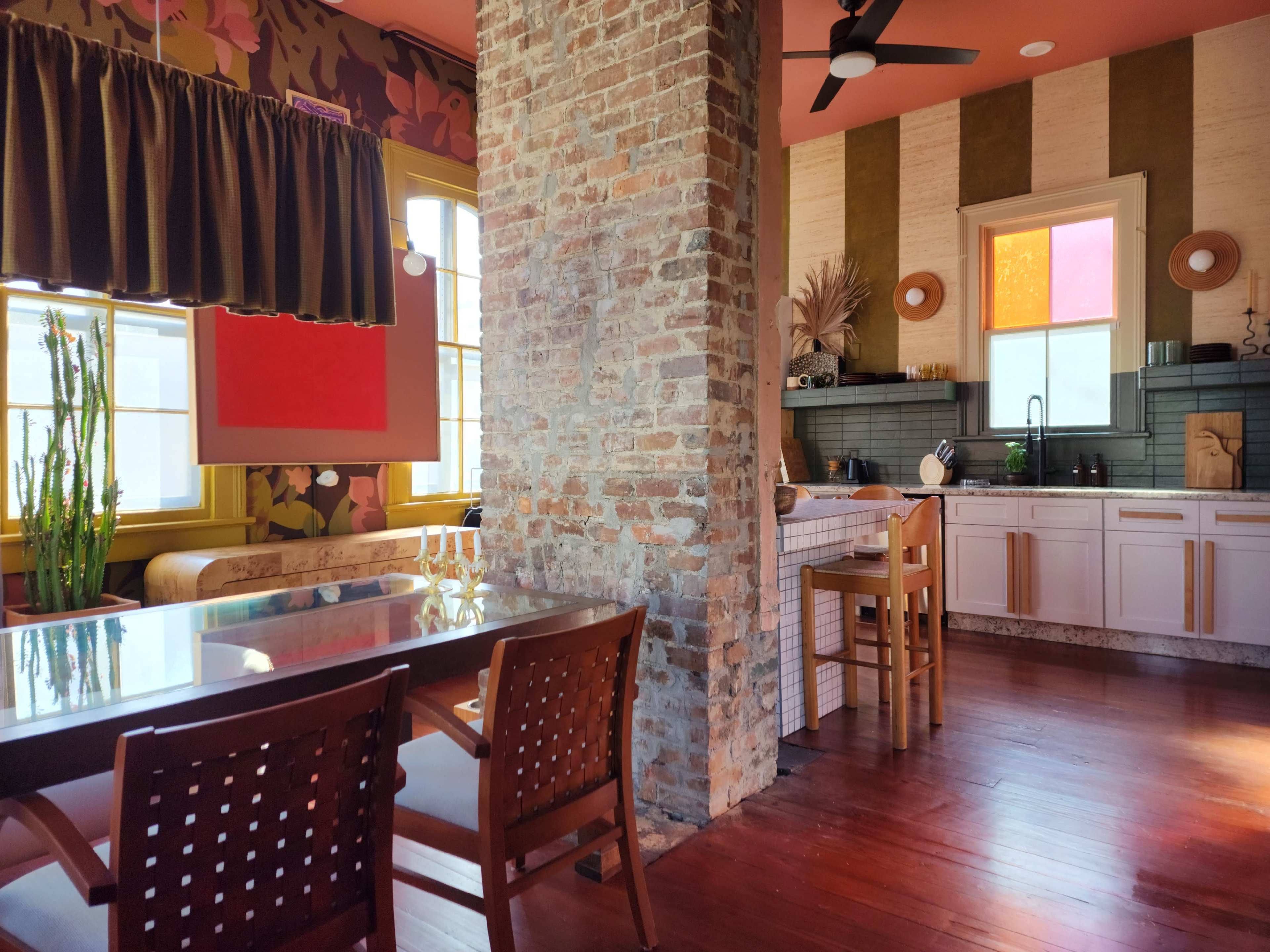 The image shows a modern kitchen and dining area with a mix of exposed brick, colorful walls, and wooden furnishings.