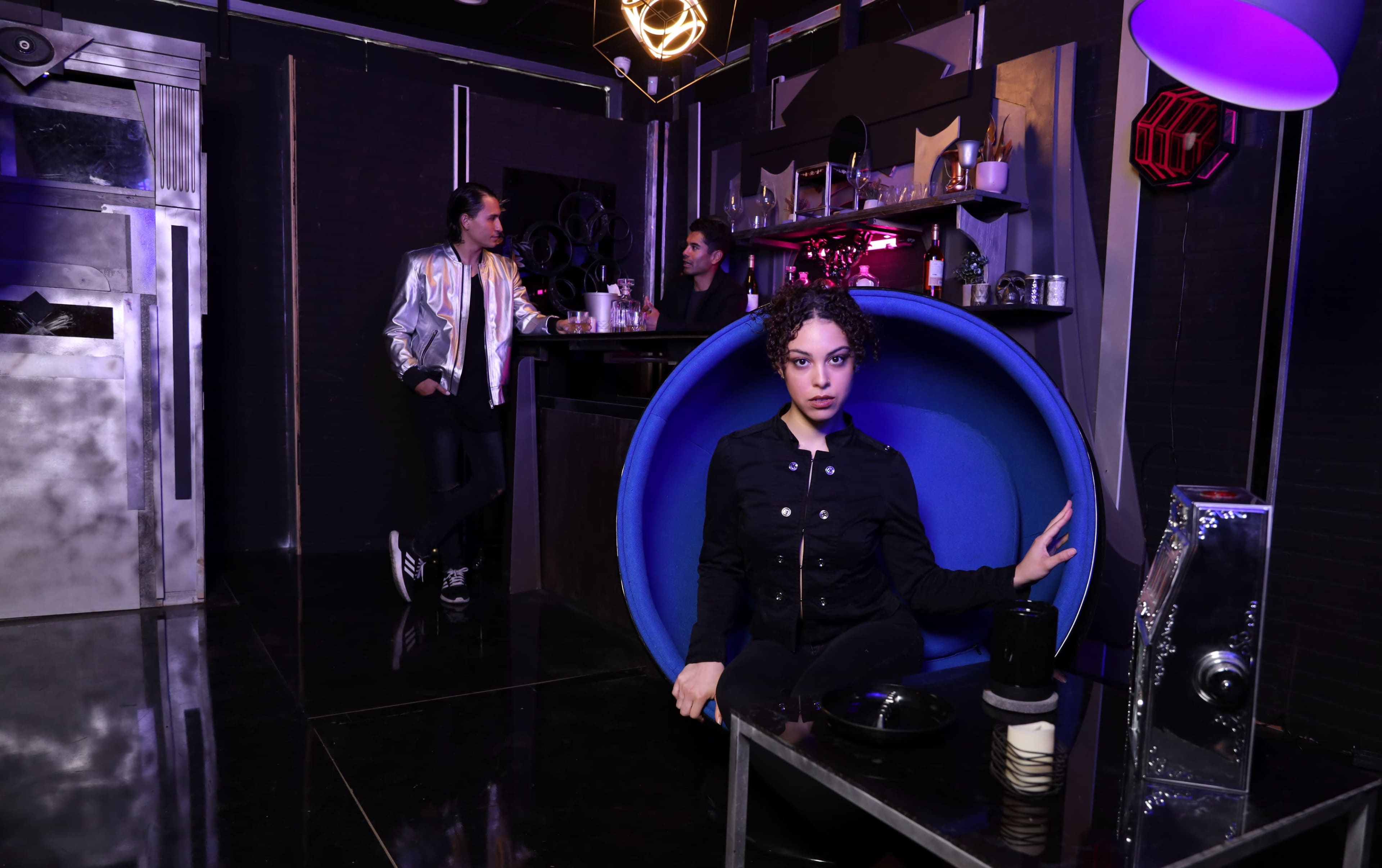 A woman sits in a blue chair in a modern bar, while two men converse behind her at the counter.
