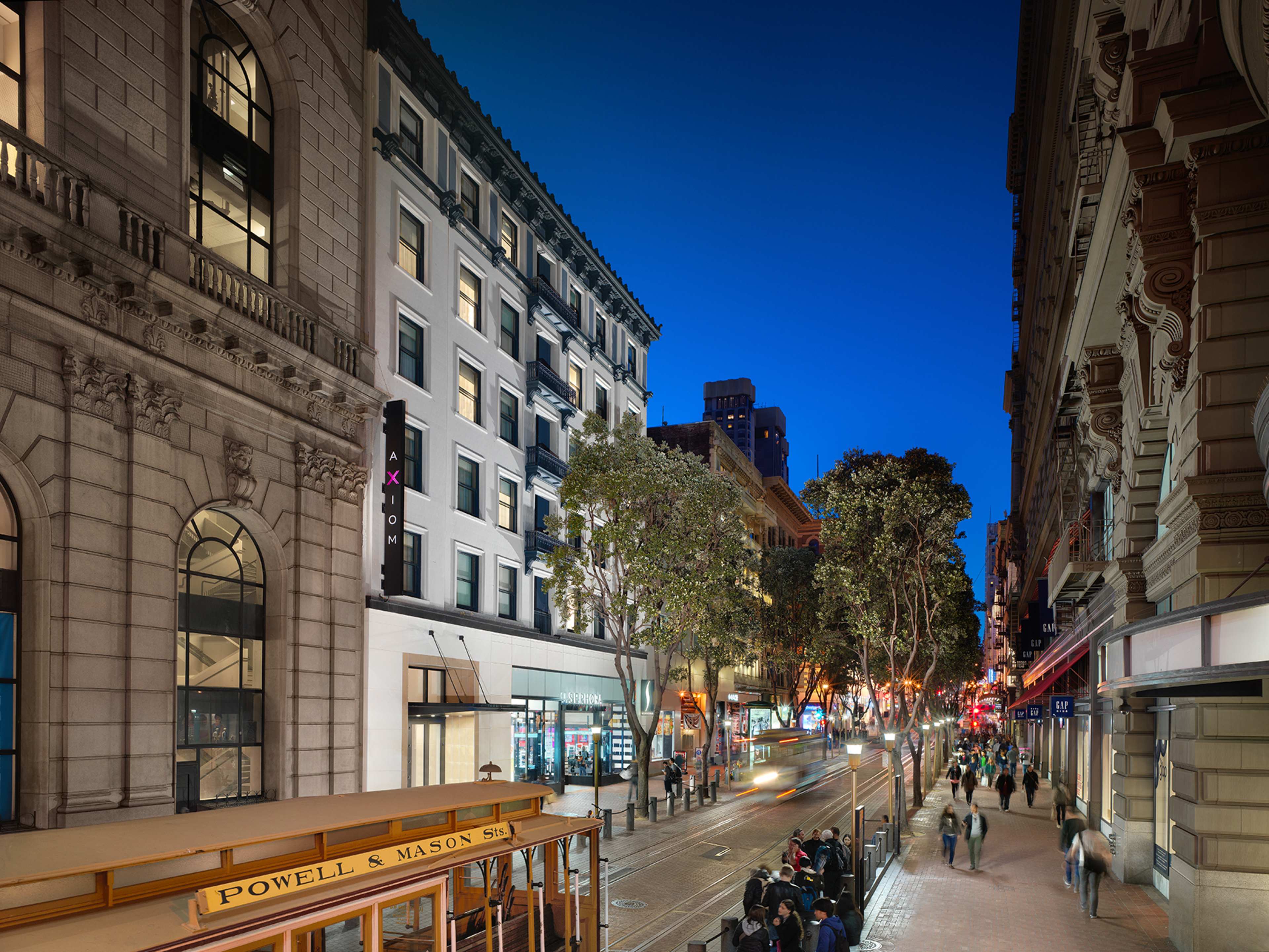 The image shows a street scene in an urban setting at dusk, featuring a cable car, lined with shops and trees along the sidewalk.