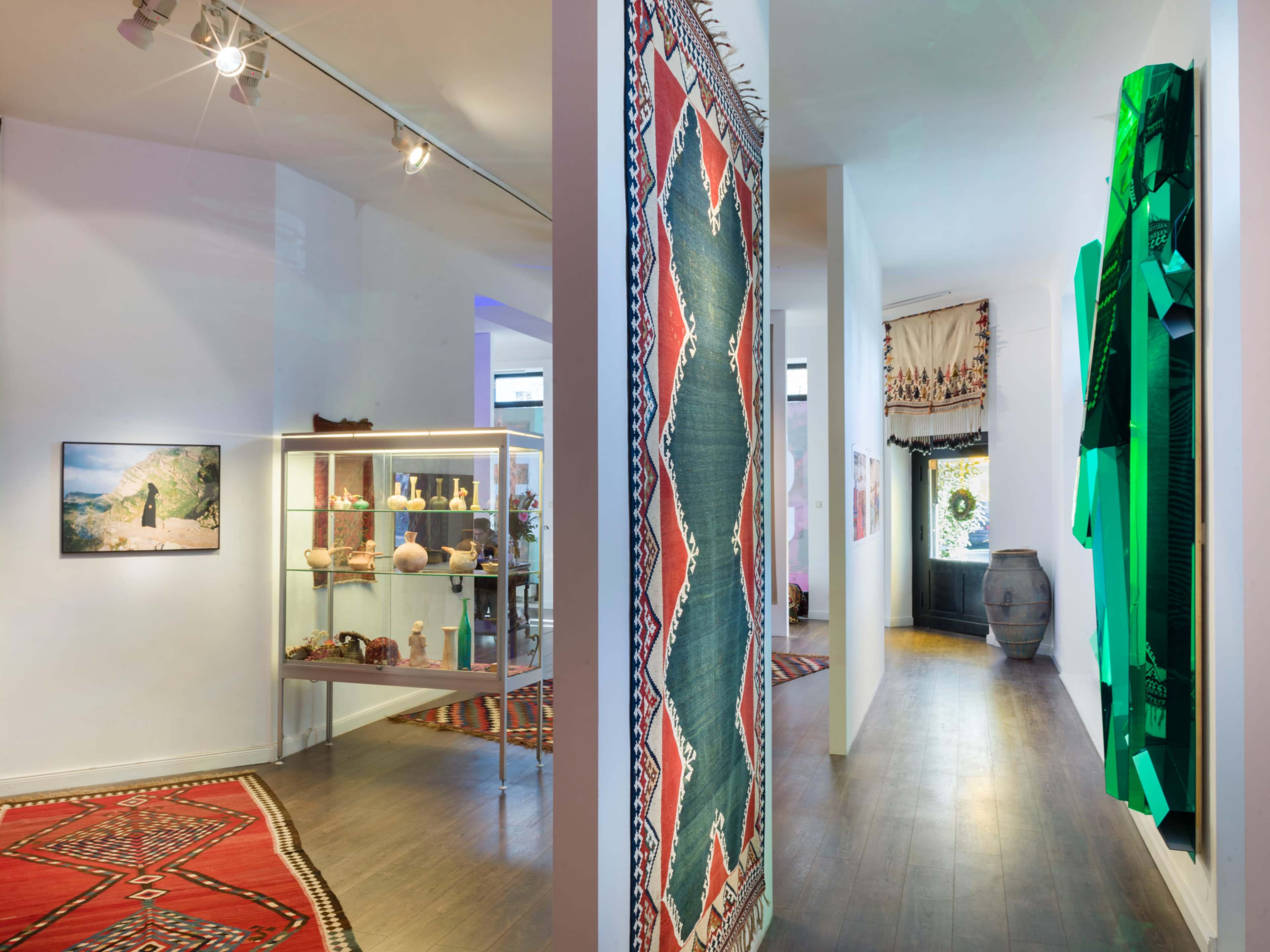 The image shows an art gallery with patterned rugs on the floor, display cases containing artifacts, and walls adorned with colorful art pieces.