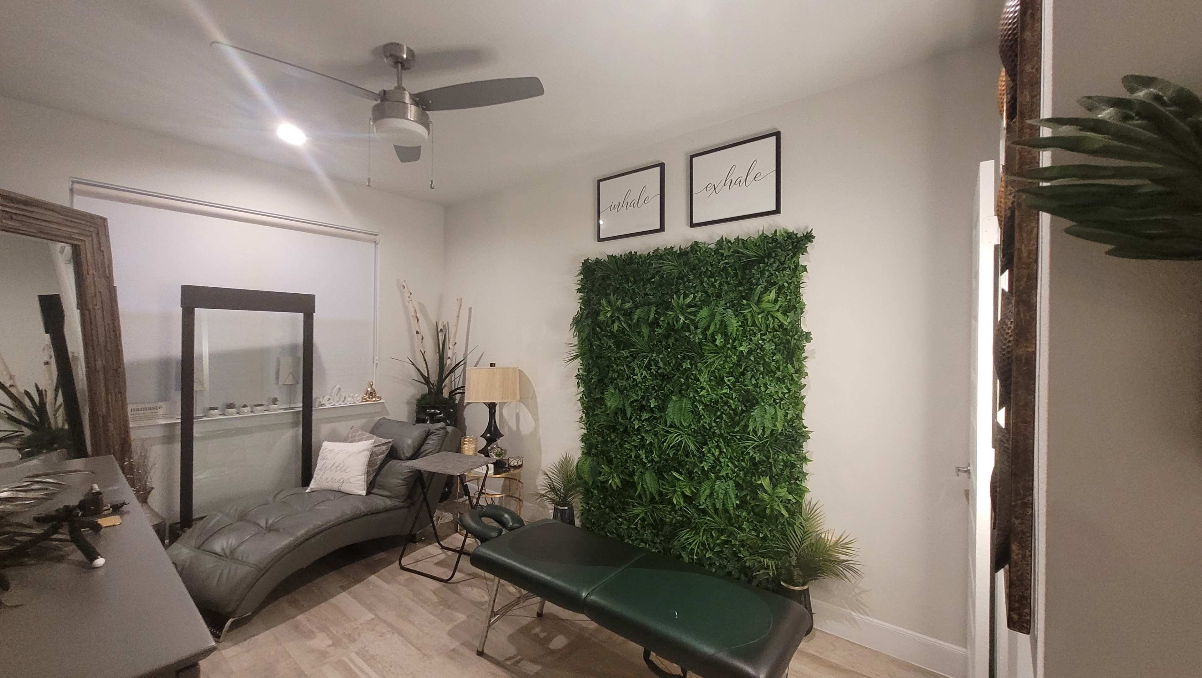 The image shows a small, well-decorated treatment room featuring a green plant wall, a treatment table, a gray couch, and decorative wall art.