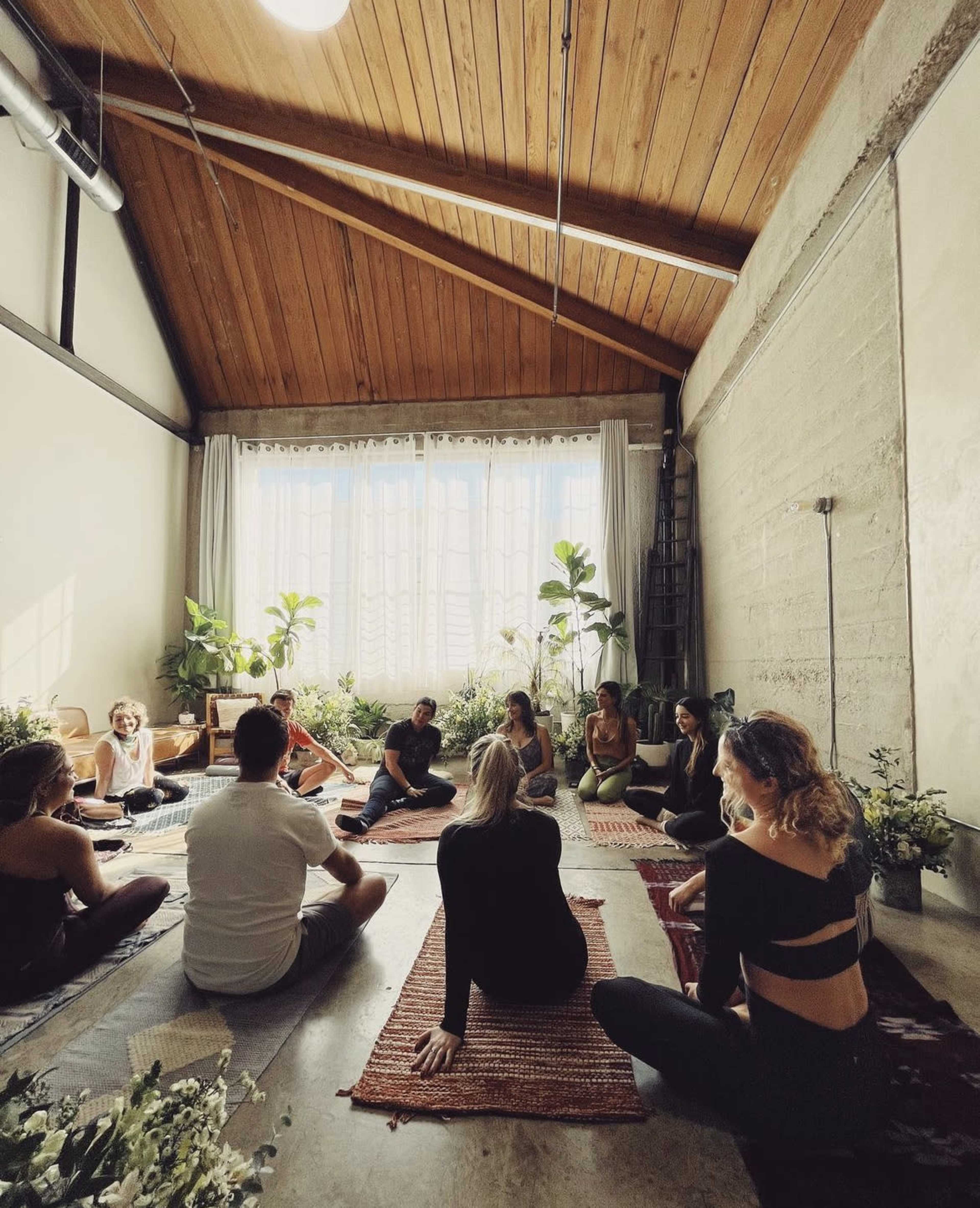 A group of people sits on mats in a spacious, well-lit room with large windows and potted plants.