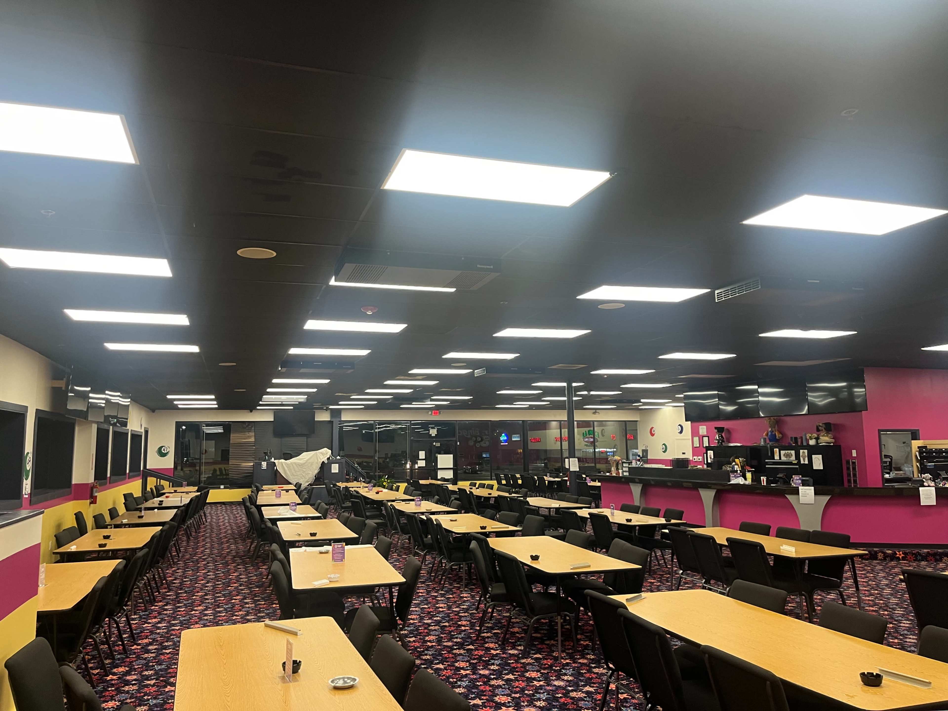A spacious dining area with tables set in rows, illuminated by bright overhead lights, and featuring pink and black decor.