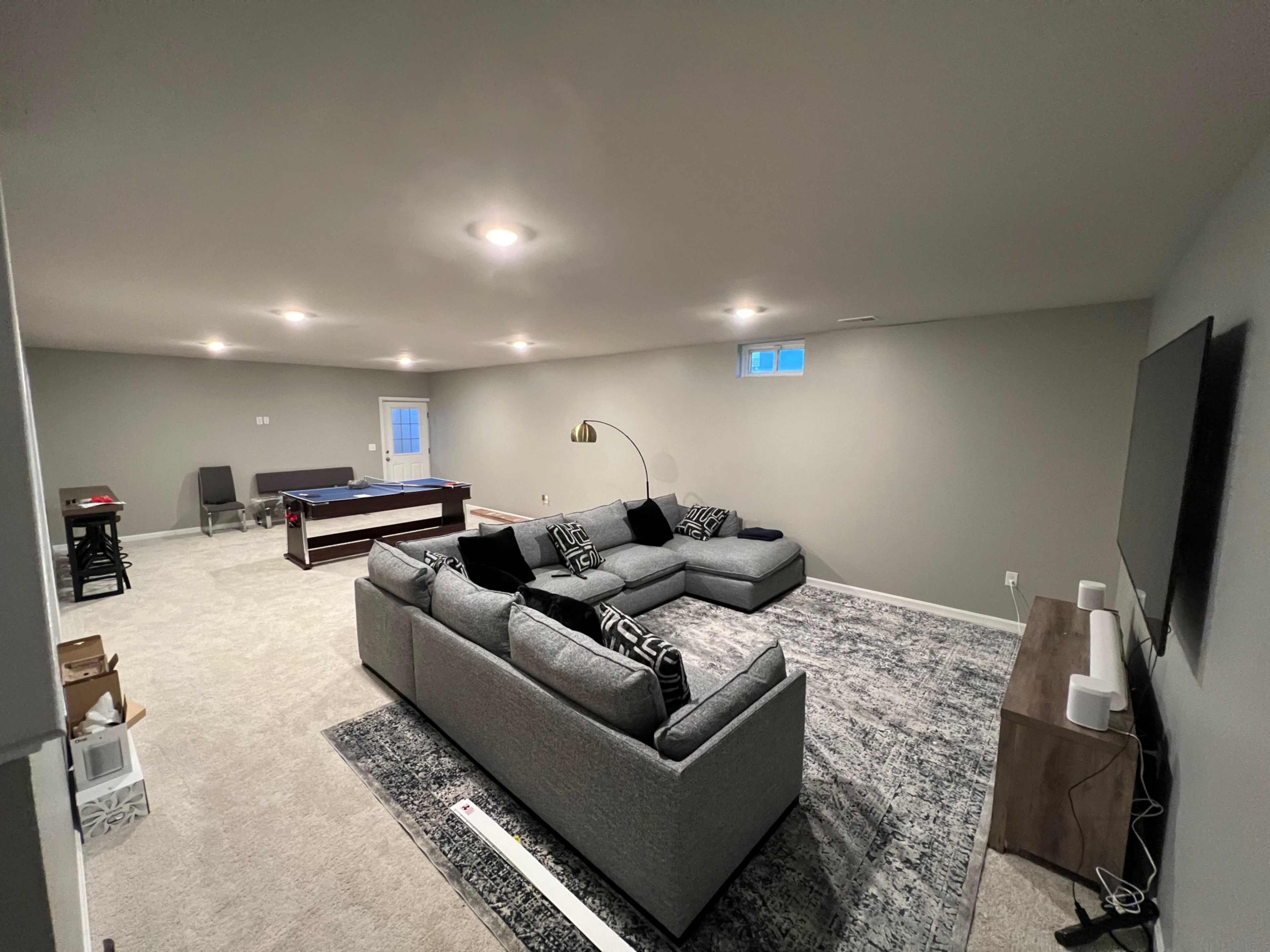 A spacious, unfinished basement features a large sectional sofa, a pool table, and several small furniture pieces against gray walls and carpet.