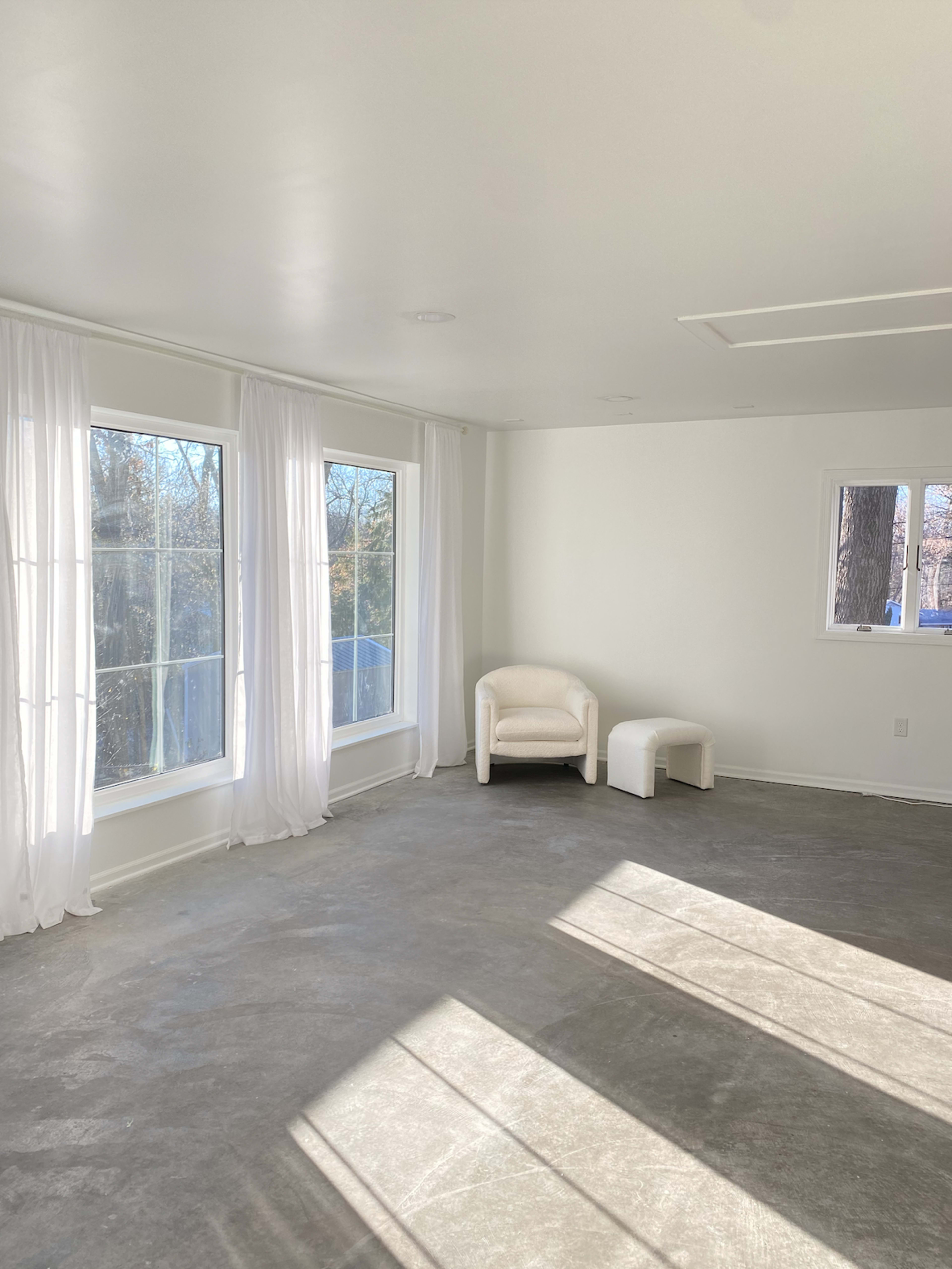 The room features large windows with sheer curtains, a white armchair, and a small stool on a bare concrete floor.
