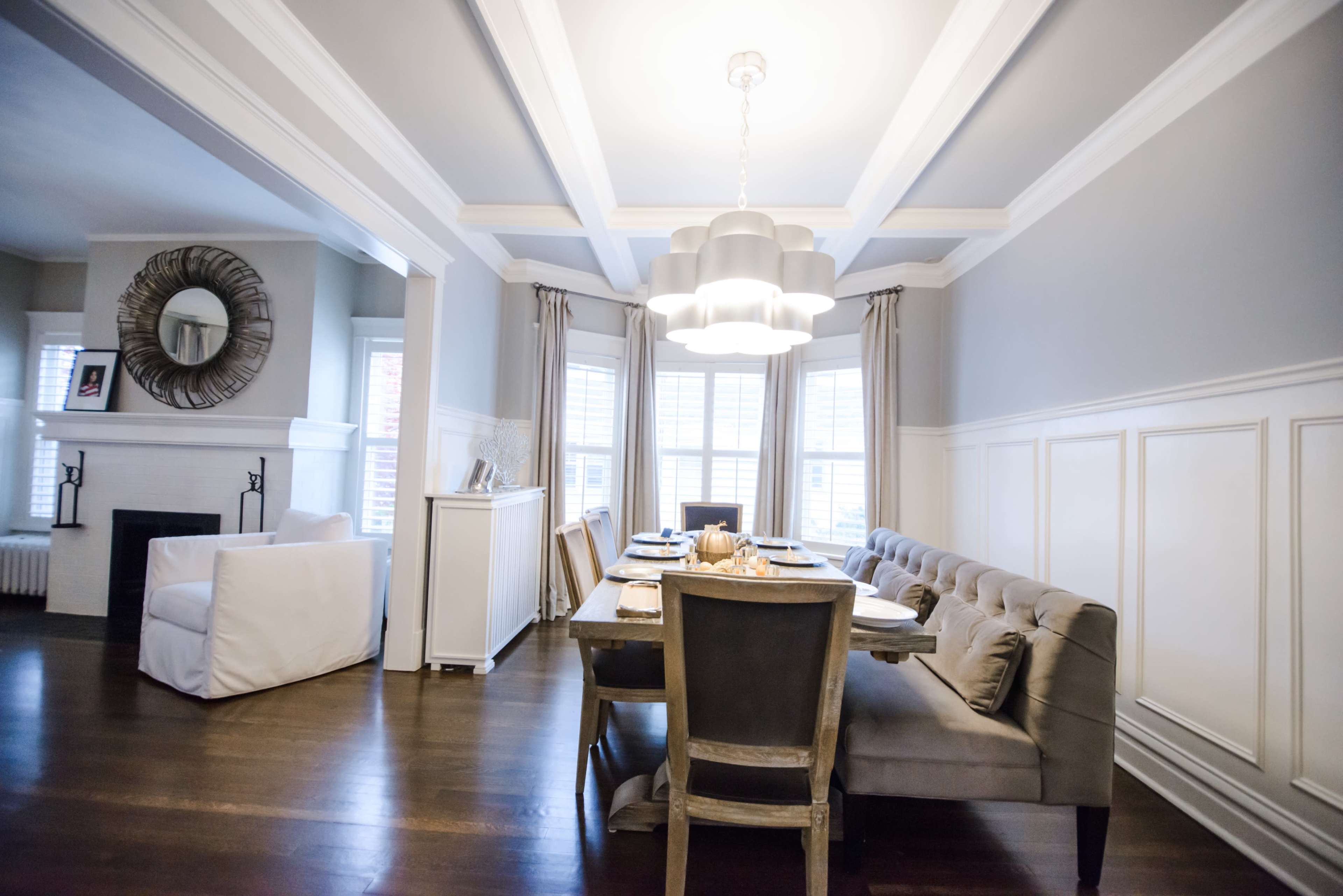 A dining area features a rectangular table set for a meal, surrounded by upholstered chairs, with large windows allowing natural light and a modern light fixture overhead.
