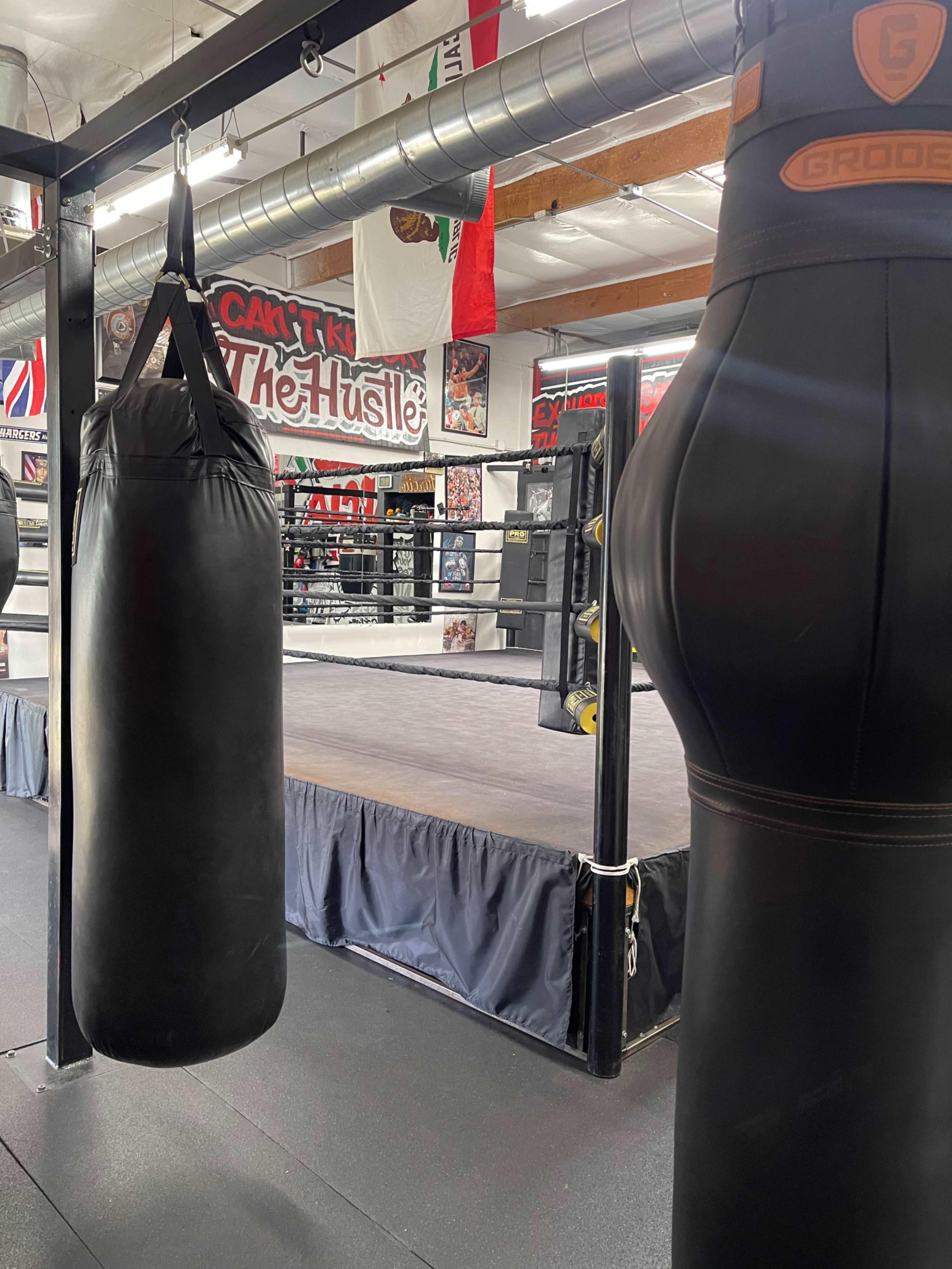A boxing gym with a heavy bag hanging in the foreground and a ring visible in the background.