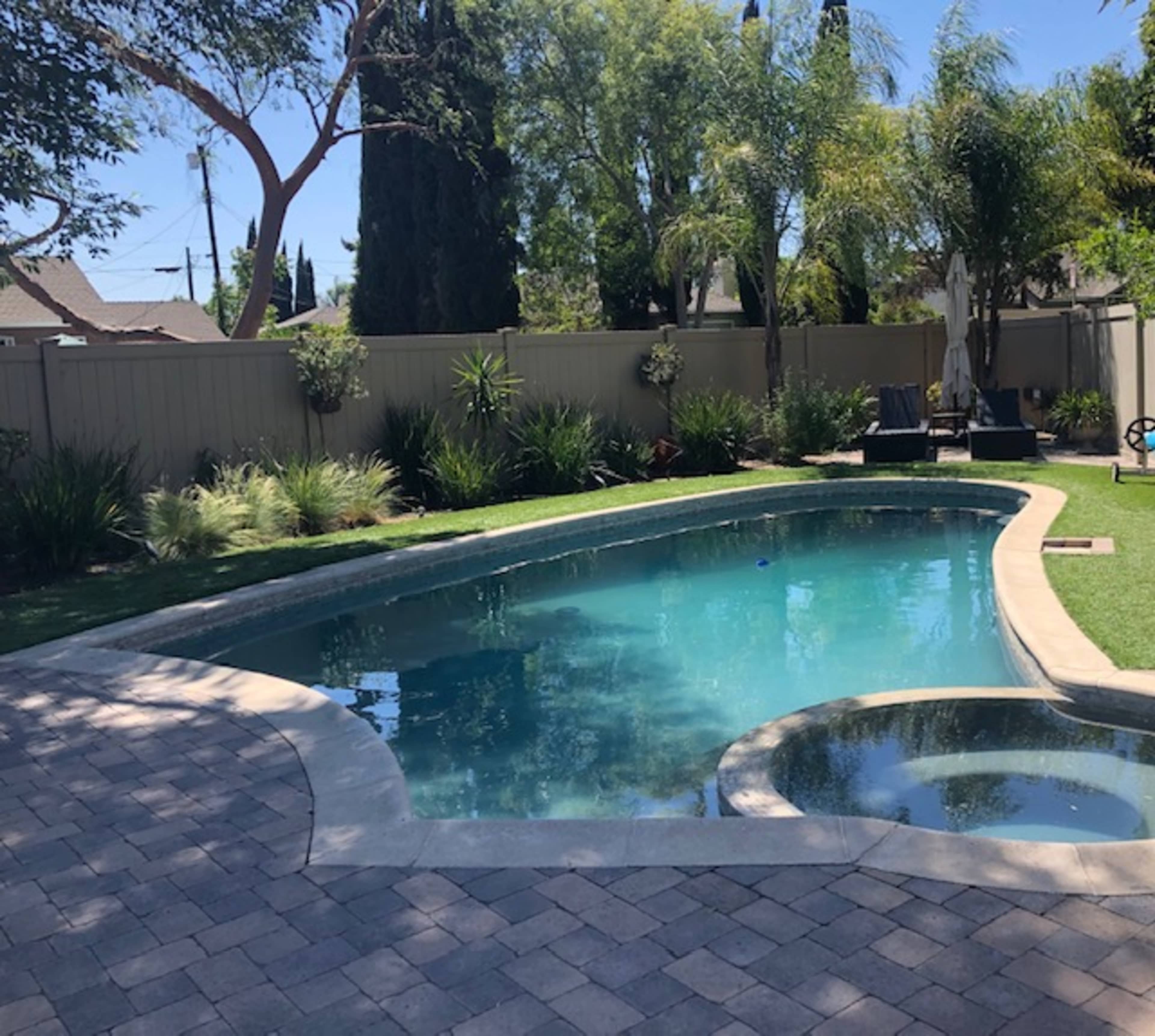 A landscaped backyard features a clear swimming pool with a spa, surrounded by greenery and paved stone.
