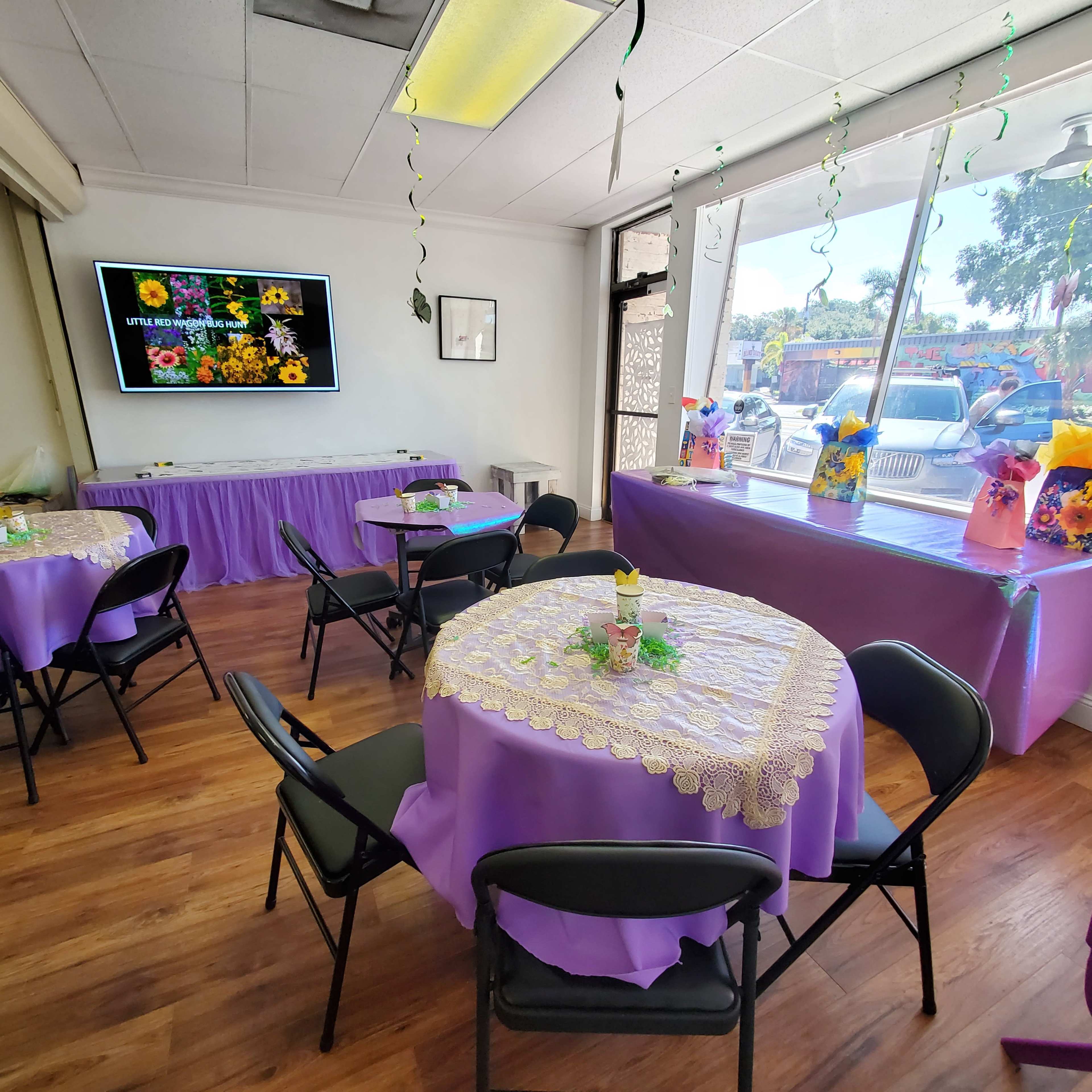 The image shows a decorated room with purple tablecloths on round tables, featuring floral centerpieces, and a large screen displaying colorful flowers on one wall.