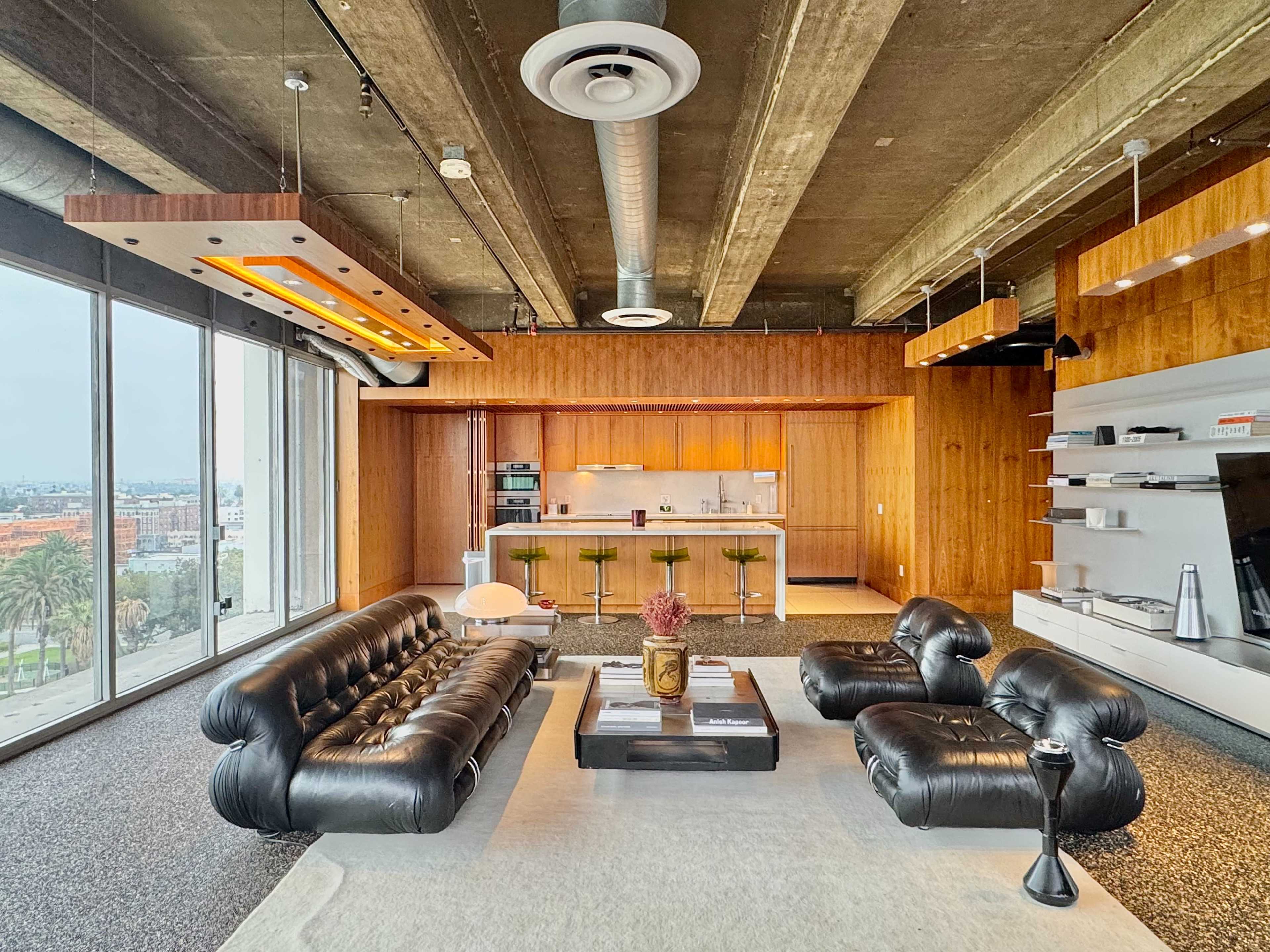 The image shows a modern living space with a black leather sofa, a coffee table, and a kitchen area featuring wooden cabinetry and bar stools.