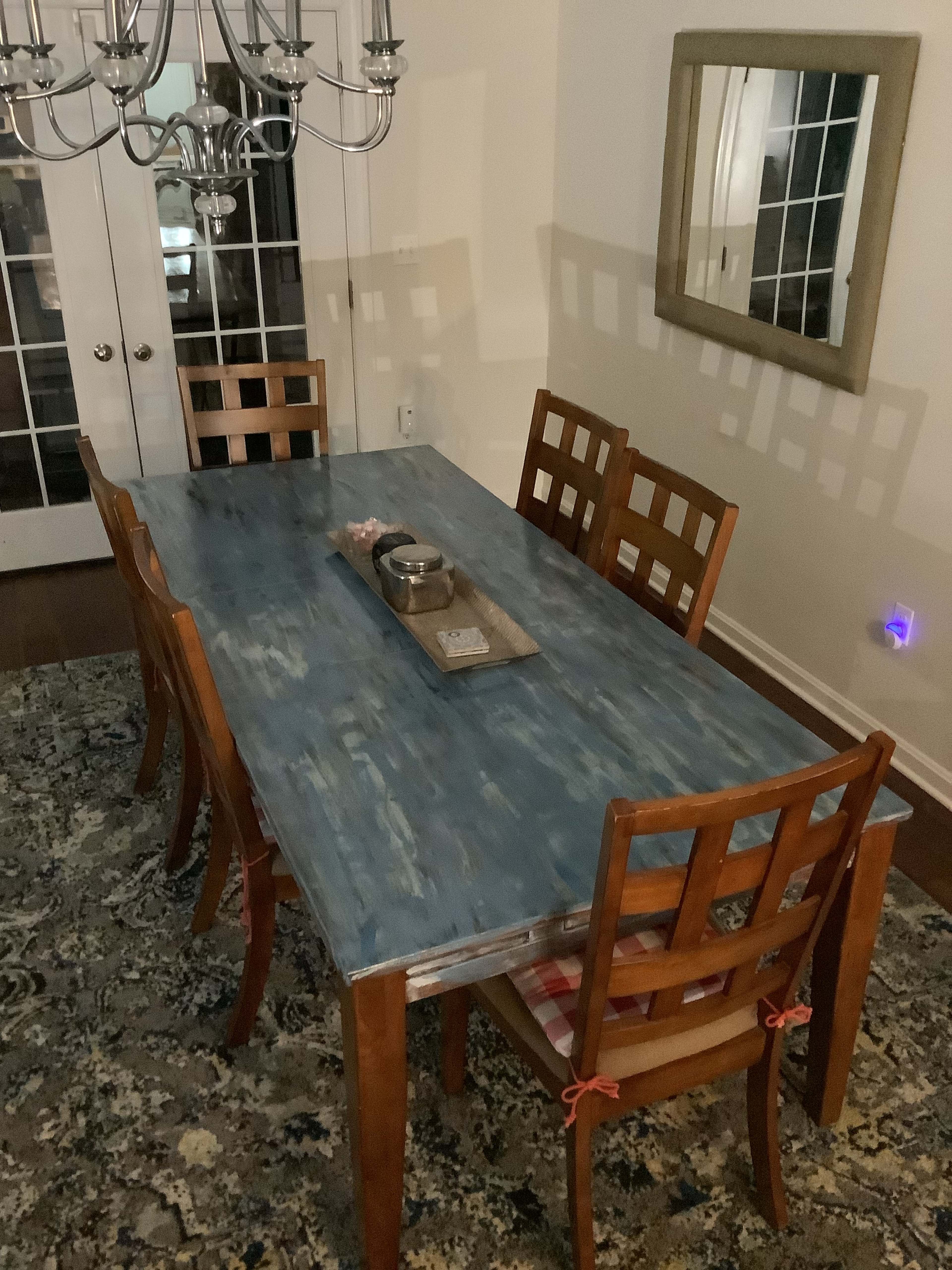 A wooden dining table with a blue surface is set in a dining area surrounded by six chairs, featuring a chandelier above and a mirror on the wall.
