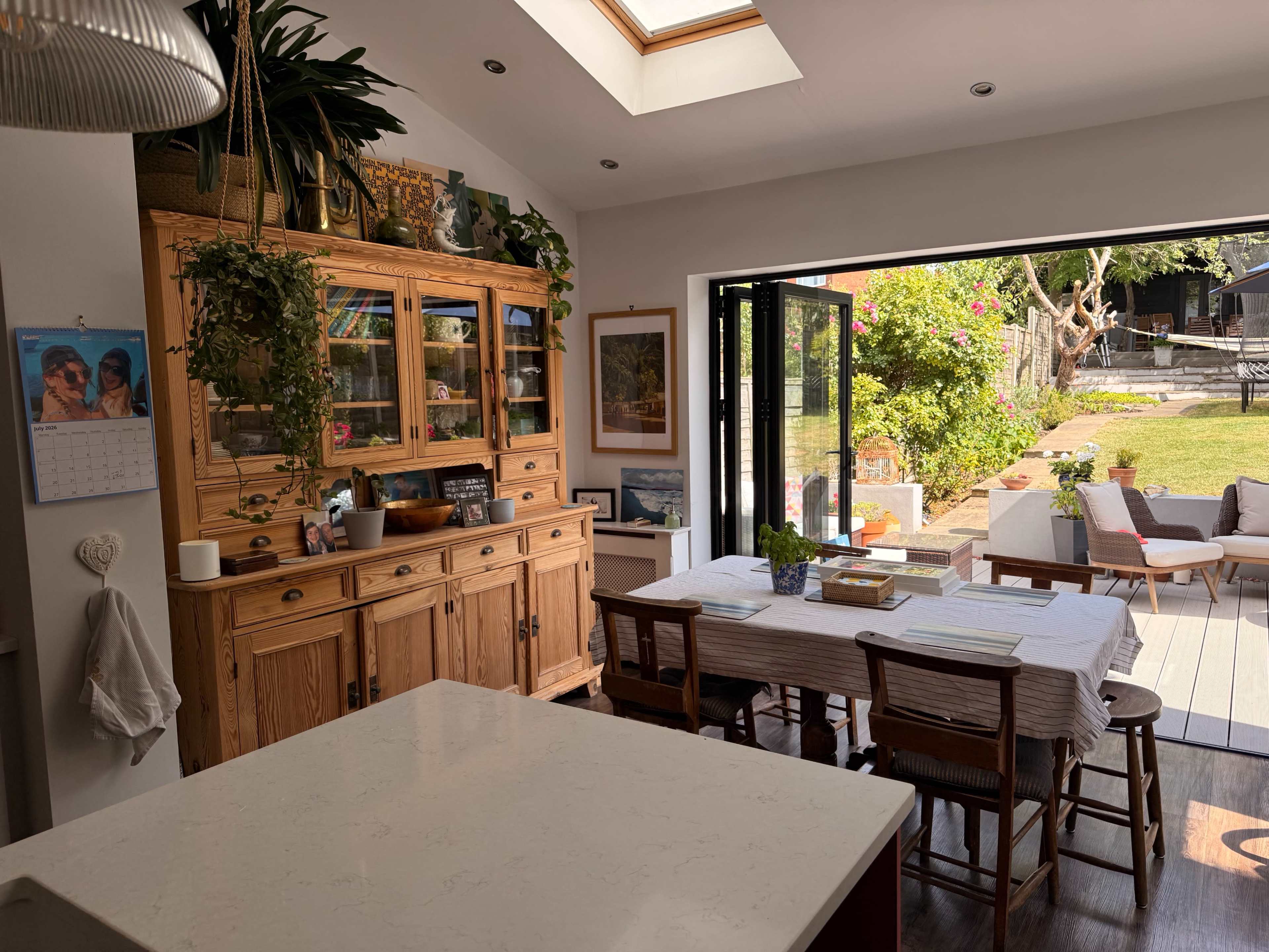 A bright kitchen features natural wood cabinetry and a dining table, with large glass doors opening to a garden area.