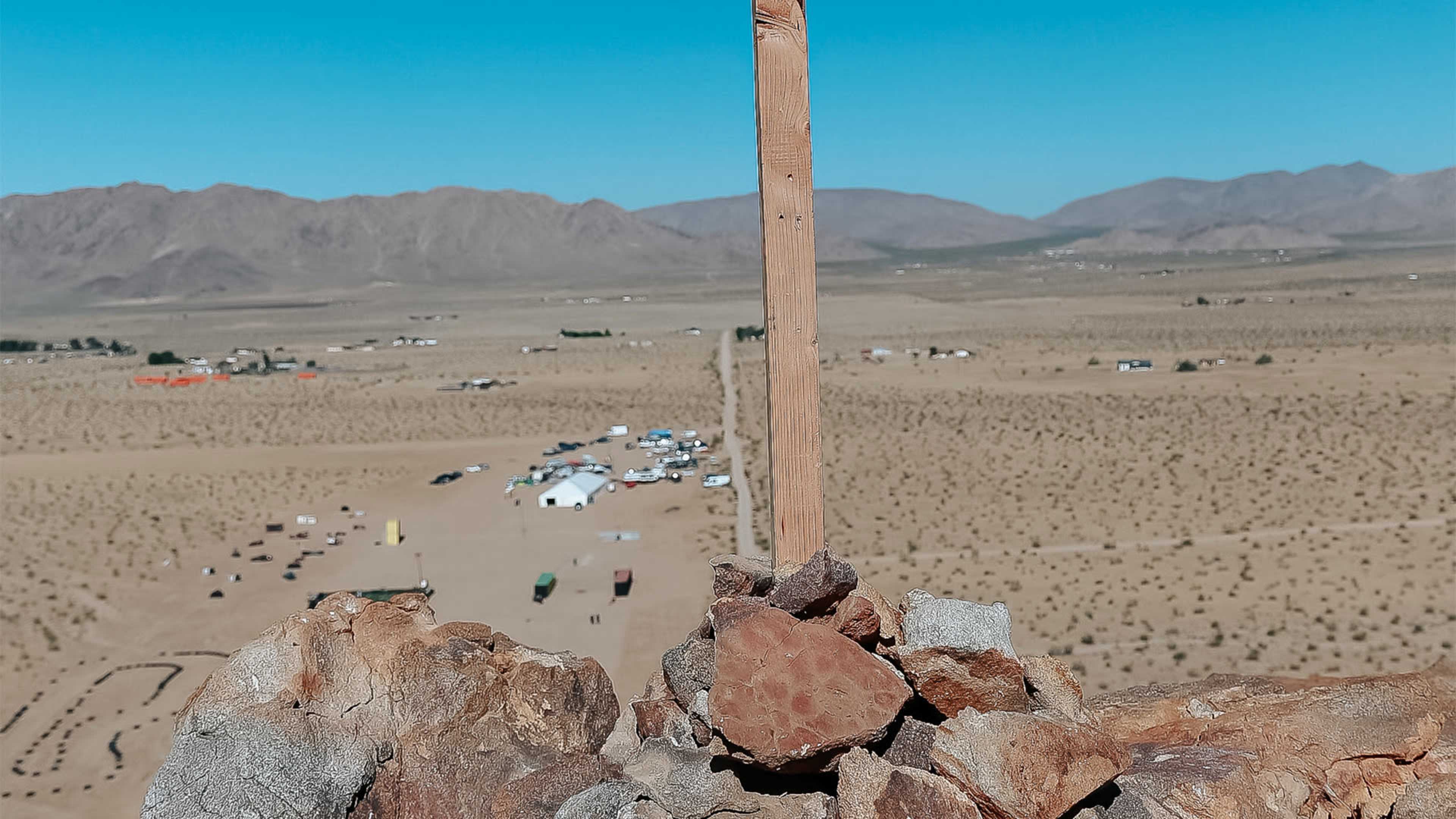 A wooden post stands atop a rock formation overlooking a sprawling desert landscape with scattered structures in the distance.