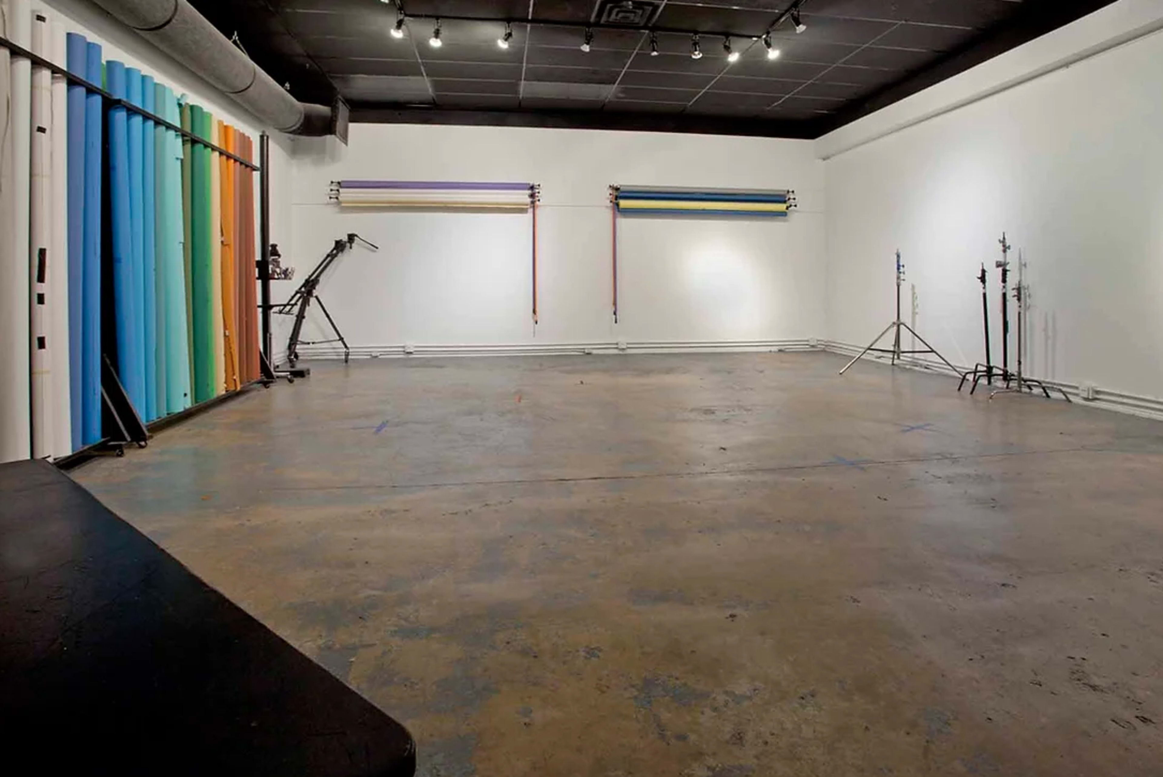 A spacious photography studio featuring colorful backdrop rolls and equipment stands on a bare concrete floor.