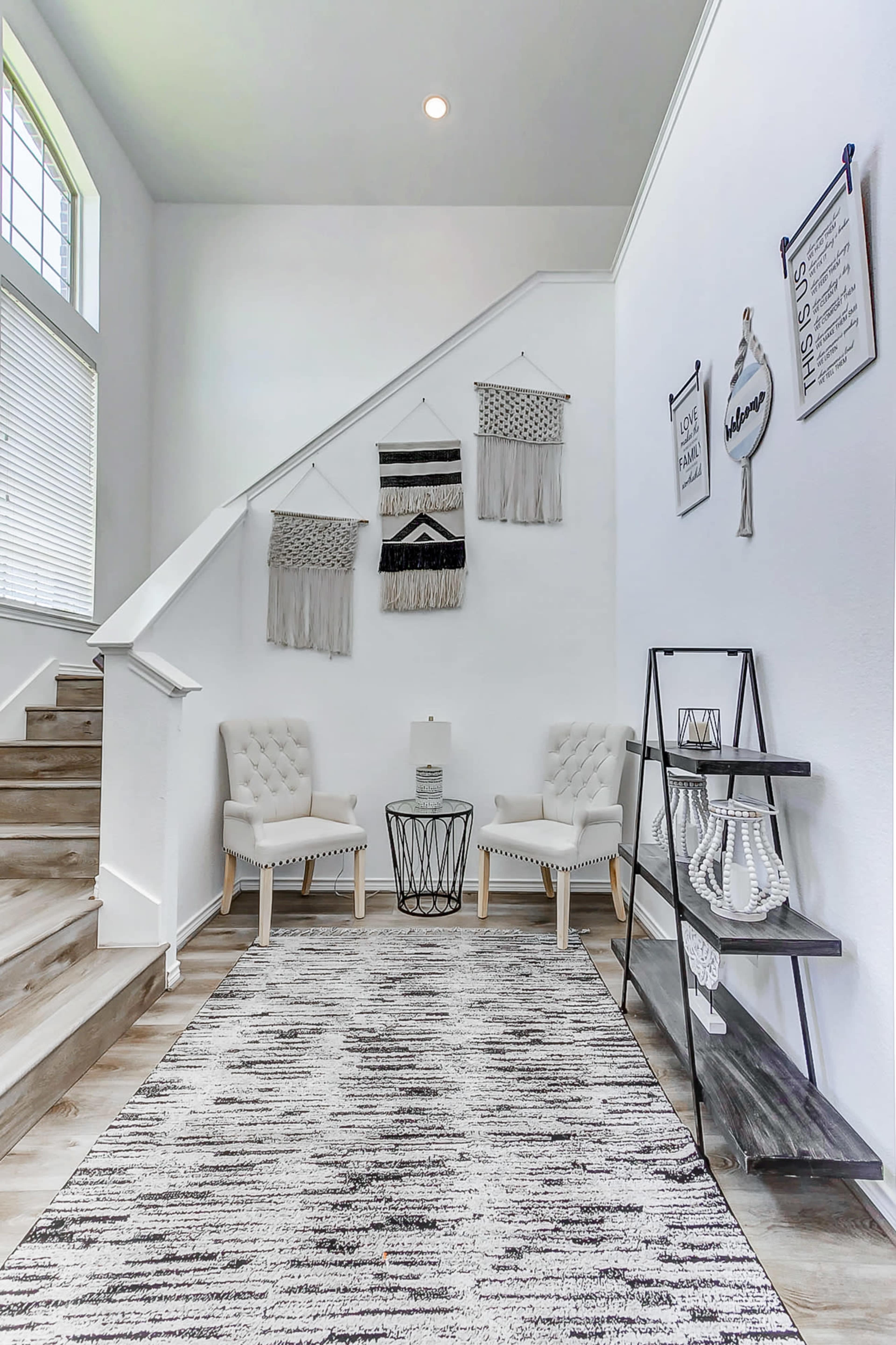A modern entryway features two upholstered chairs, a circular table, and decorative wall hangings beside a staircase.